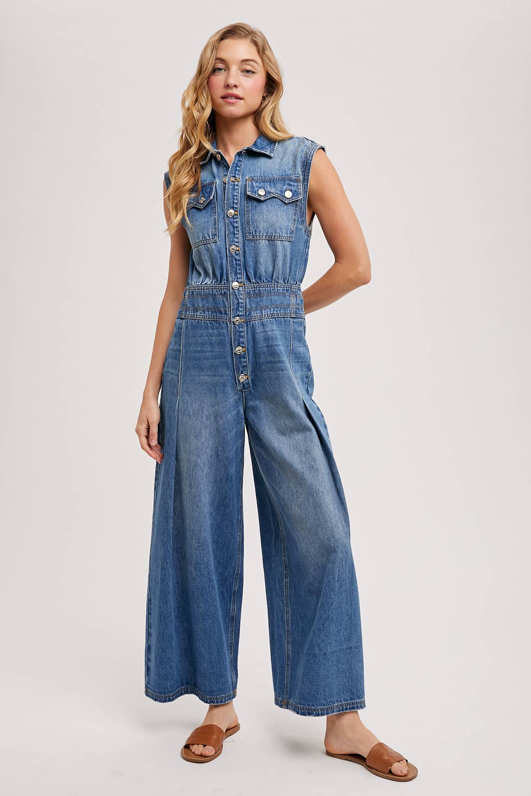 Sleeveless denim jumpsuit with collared button-front, chest flap pockets, and wide-leg palazzo silhouette, shown on model with slide sandals.
