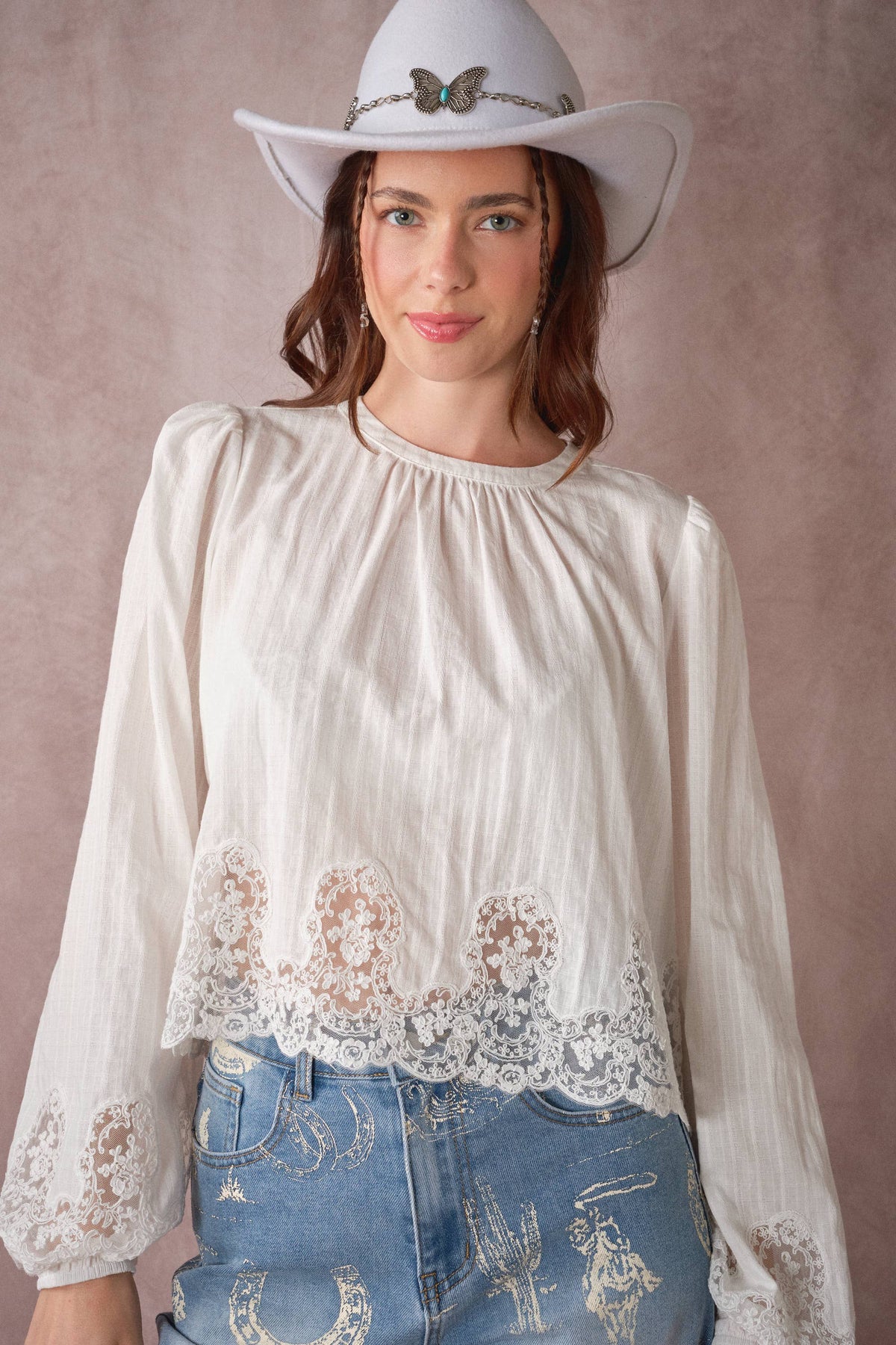 A woman wearing a white long-sleeve blouse with lace trim at the hem and cuffs, paired with blue jeans and a white cowboy hat, posed against a textured mauve-pink studio backdrop.