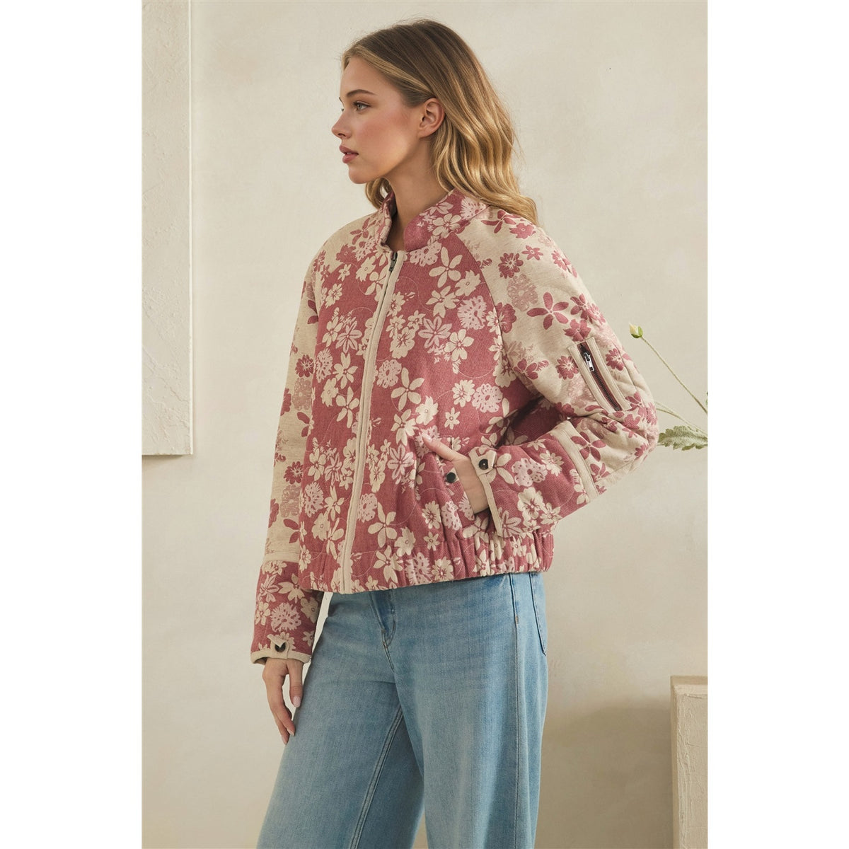 A woman wearing a pink and beige floral print bomber jacket with light blue jeans, standing in a neutral indoor setting.