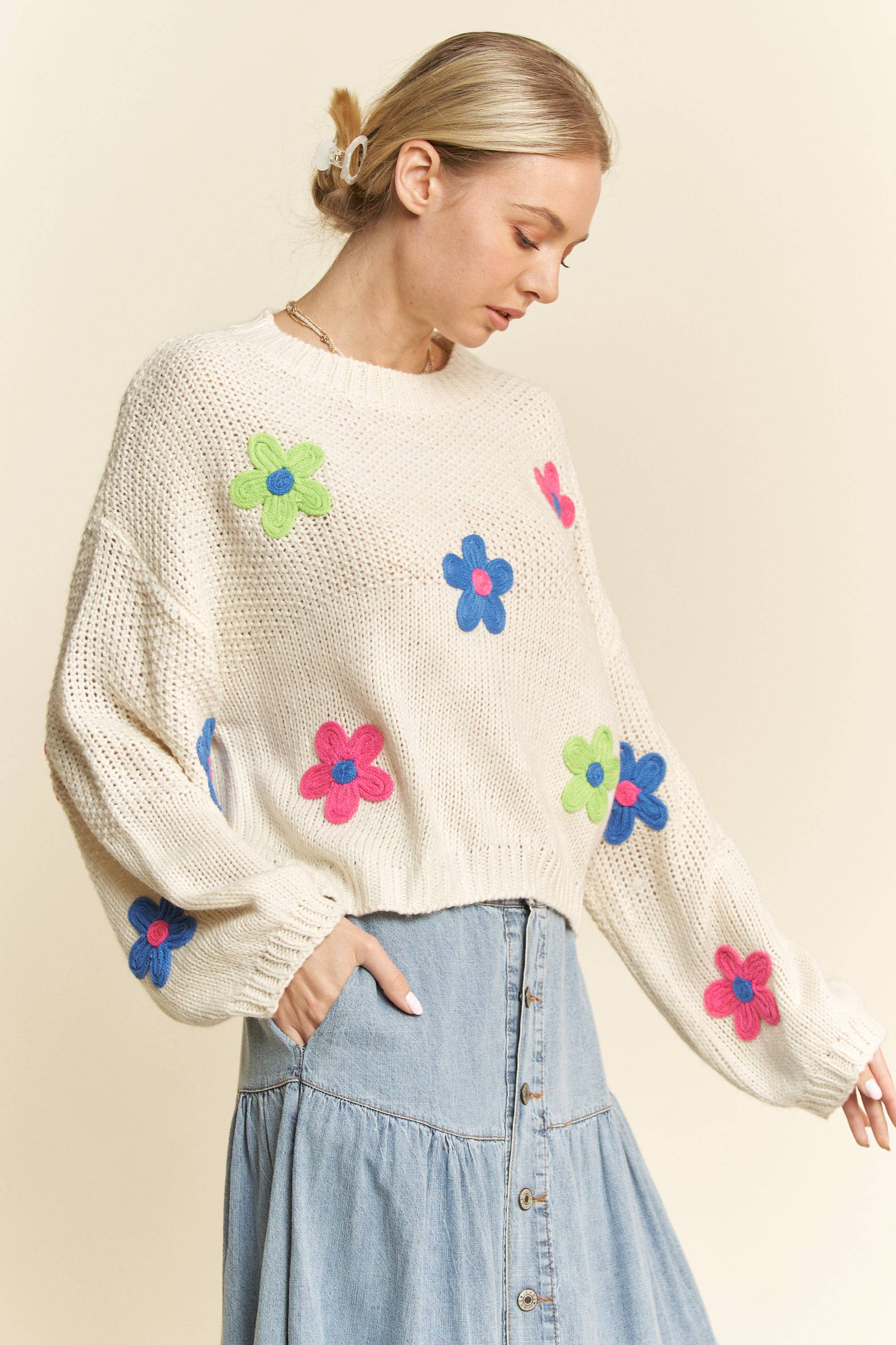 Cream knit sweater with colorful 3D floral appliqués in pink, blue, and green, styled with a button-front light denim skirt.