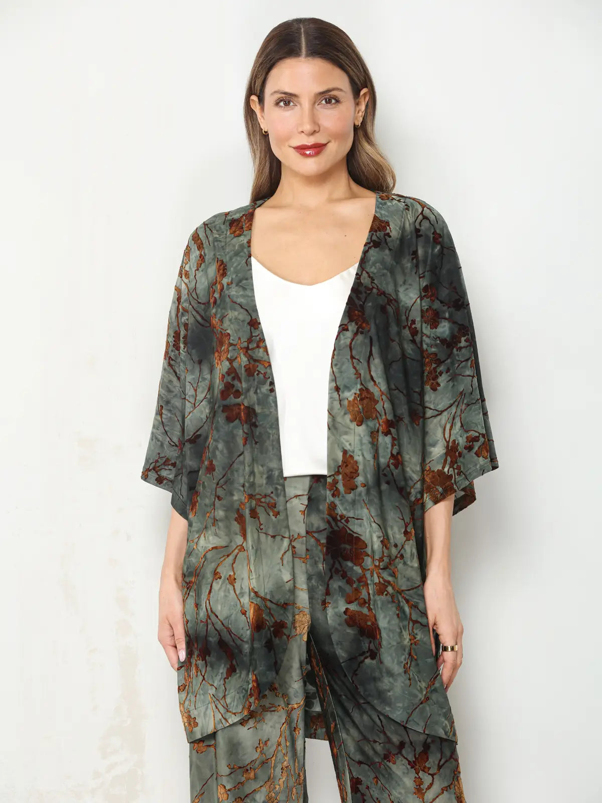 A woman modeling a matching two-piece lounge set featuring a kimono cardigan and pants in a green and brown abstract floral print, worn over a white tank top against a light gray wall.