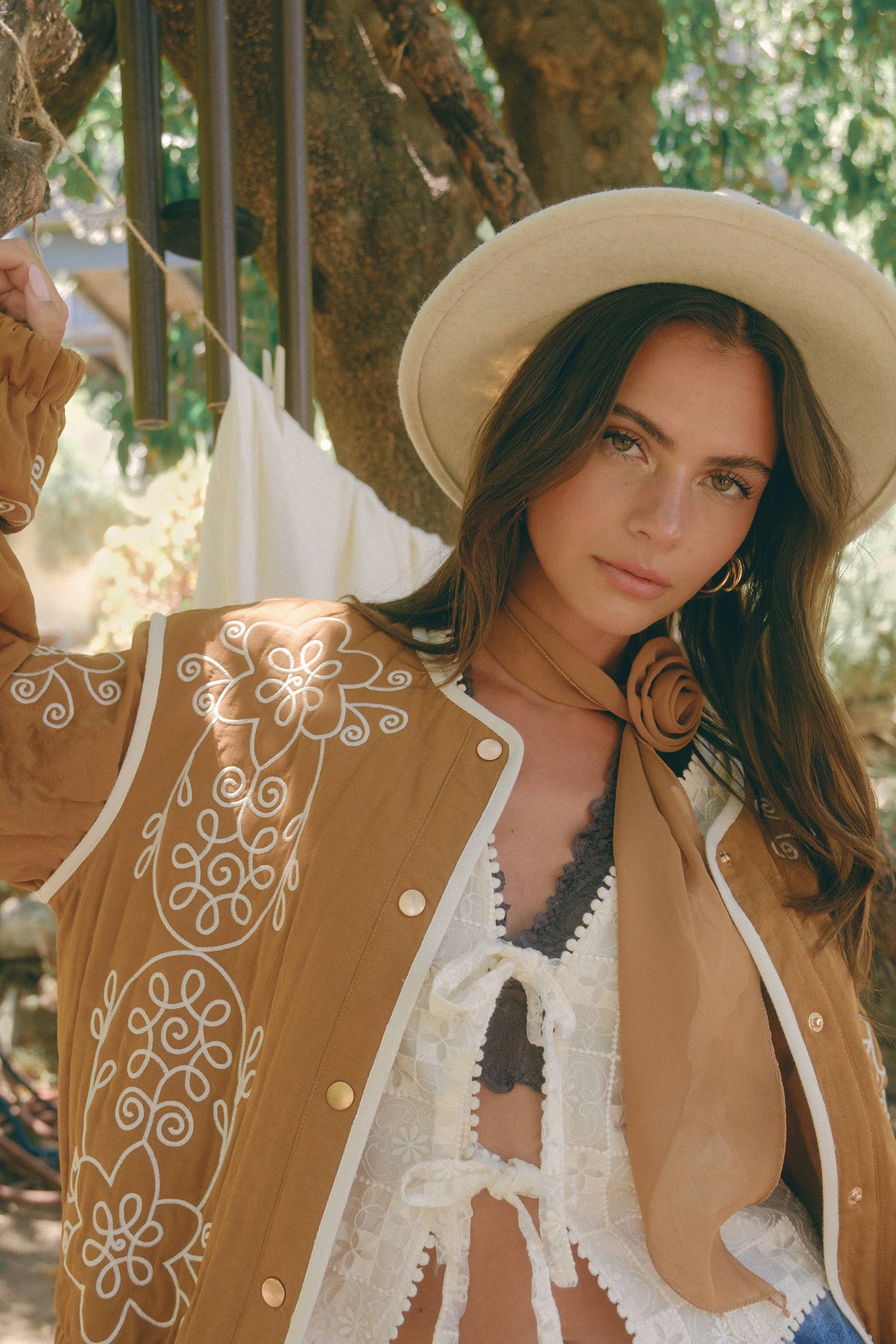 Boho caramel-brown jacket with cream floral swirl embroidery, ivory piping, and snap buttons, styled over a lace tie-front top with a scarf and felt hat outdoors.