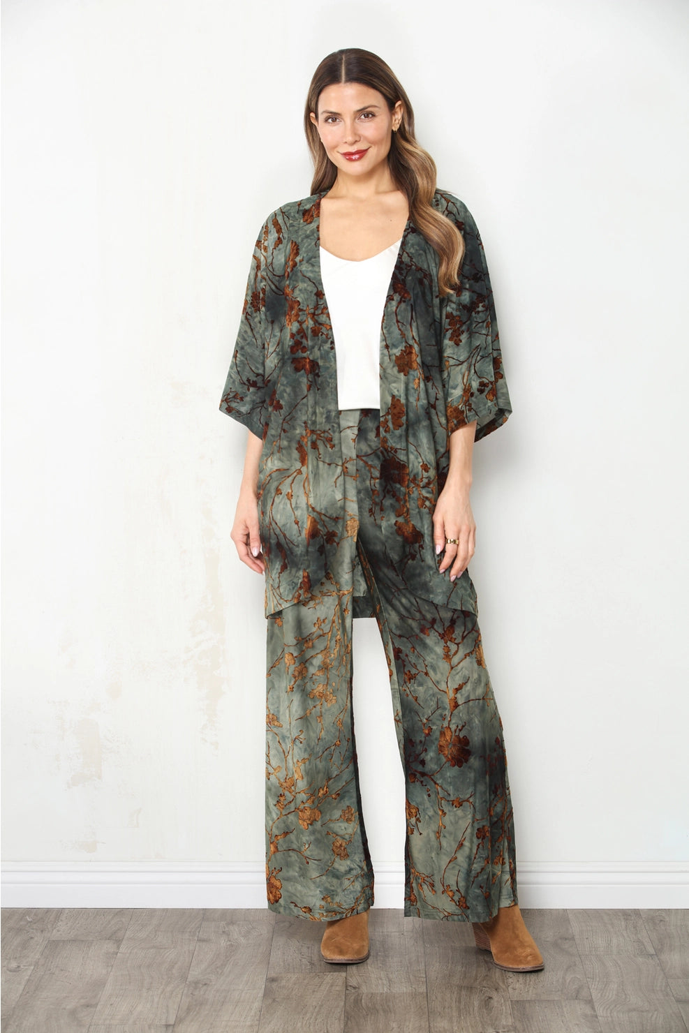 A woman modeling a matching two-piece lounge set featuring a kimono jacket and wide-leg pants in a green and brown abstract print, styled over a white top and brown boots against a white wall.