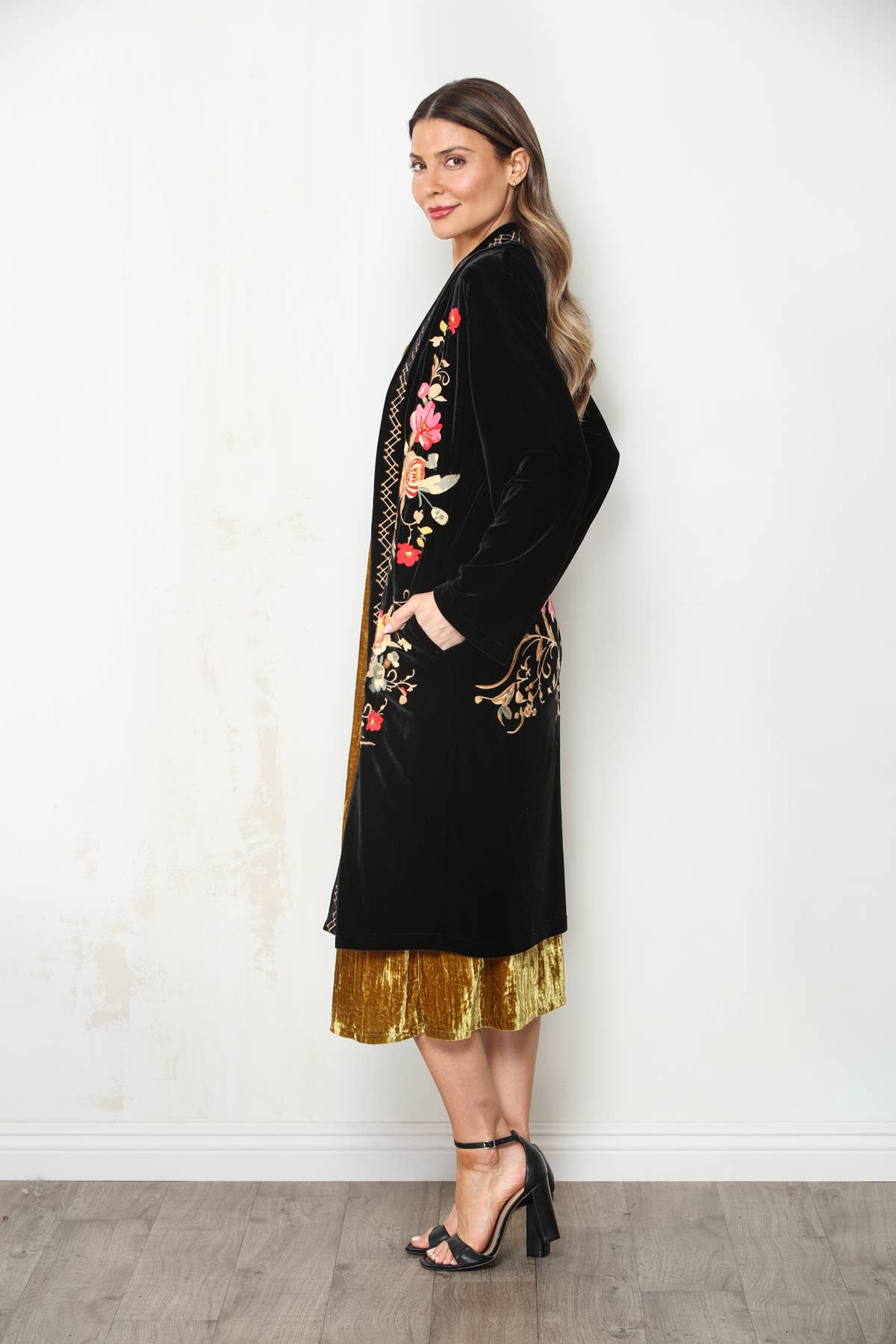 Side view of long black velvet duster with floral embroidery, long sleeves and side pocket, worn over a gold dress.