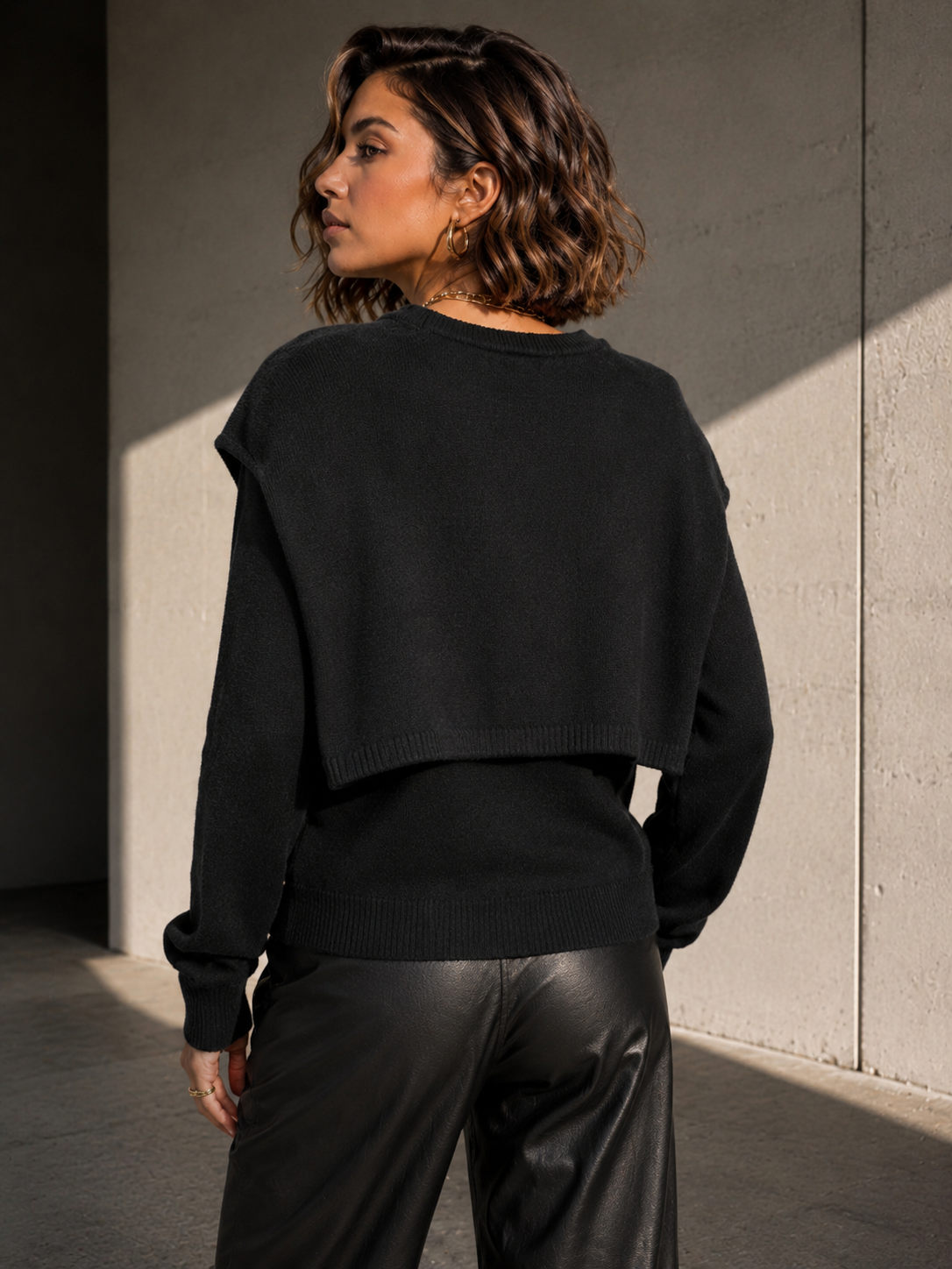 A woman wearing a black cropped knit sweater and black leather pants, photographed from behind in a sunlit concrete corridor.