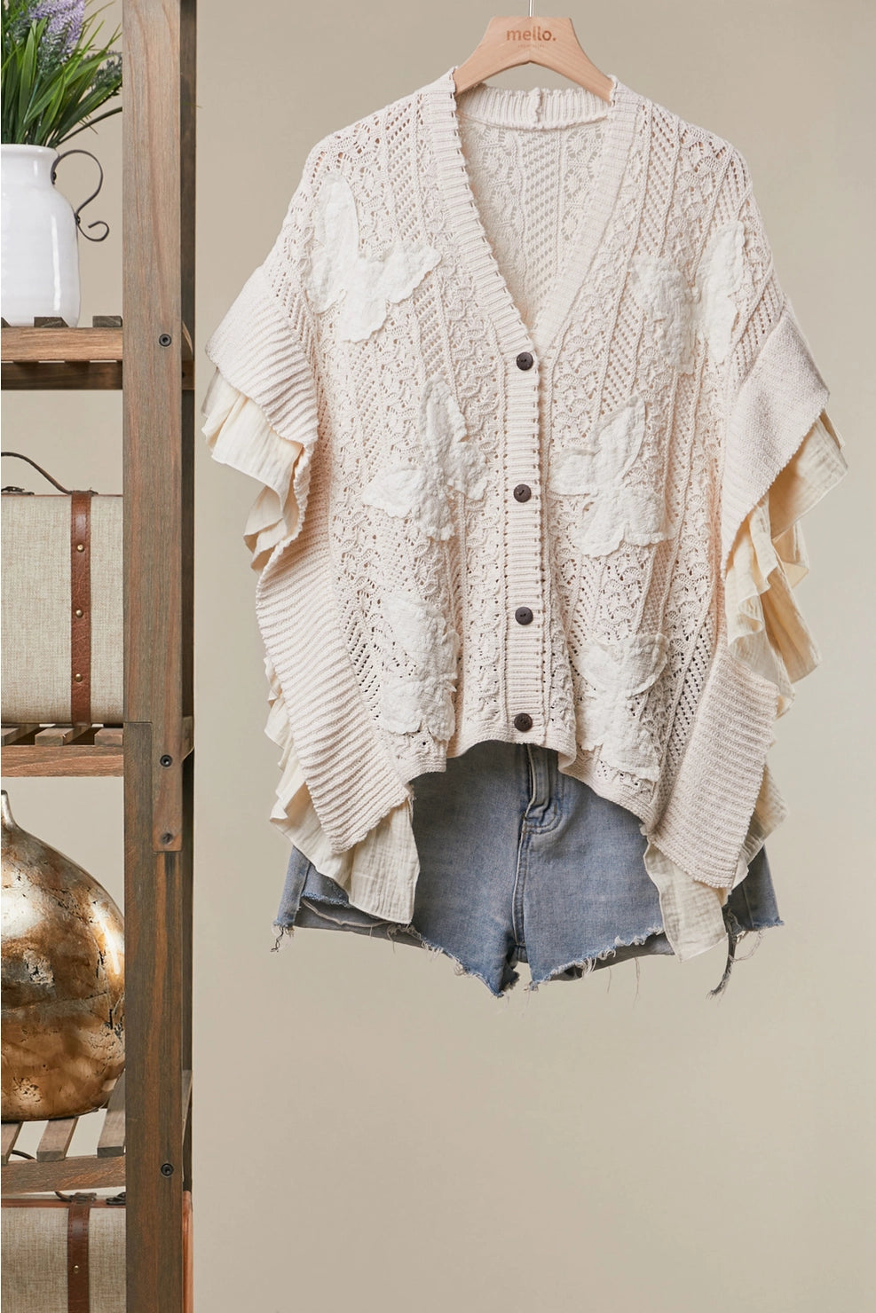 Butterfly Bloom Cardigan Cream-colored textured knit cardigan with ruffle details and button-front closure, styled over denim shorts on a hanger against a beige wall.