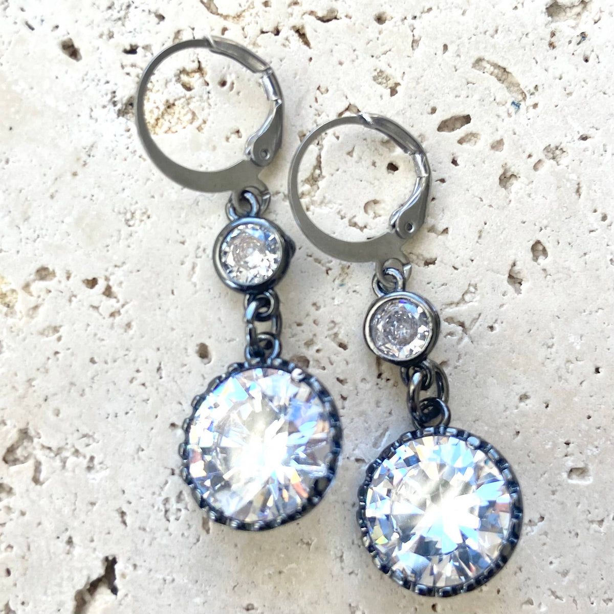 A pair of drop earrings with gunmetal leverback hoops, small clear round stones, and large faceted iridescent clear stones in gunmetal settings, displayed on a light textured stone surface.