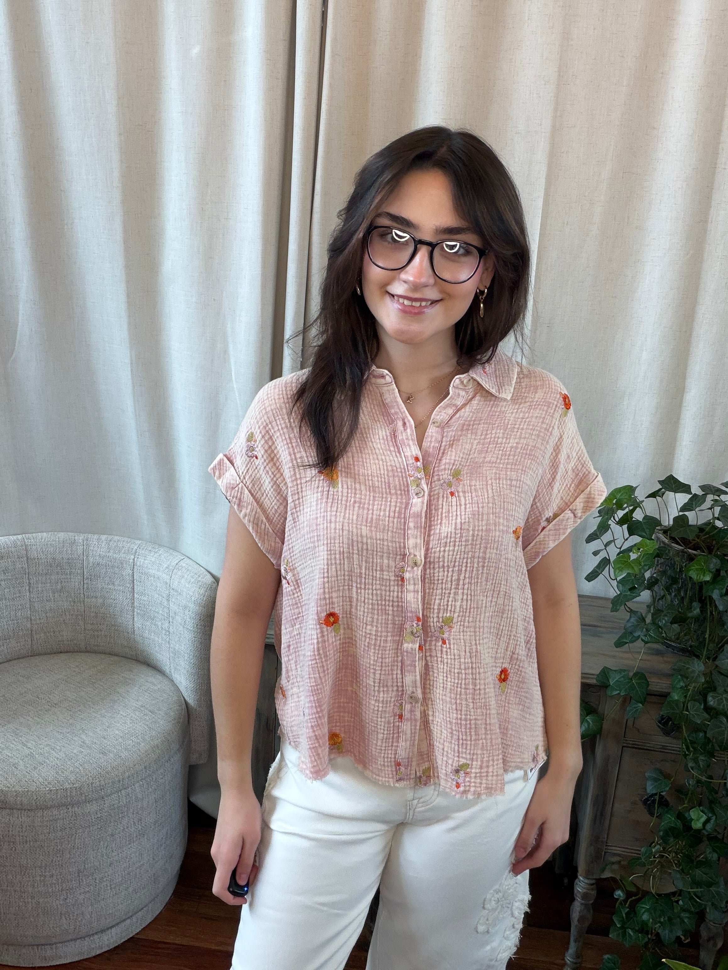 Woman wearing a pink checkered blouse with white pants standing in a room with curtains and a chair.