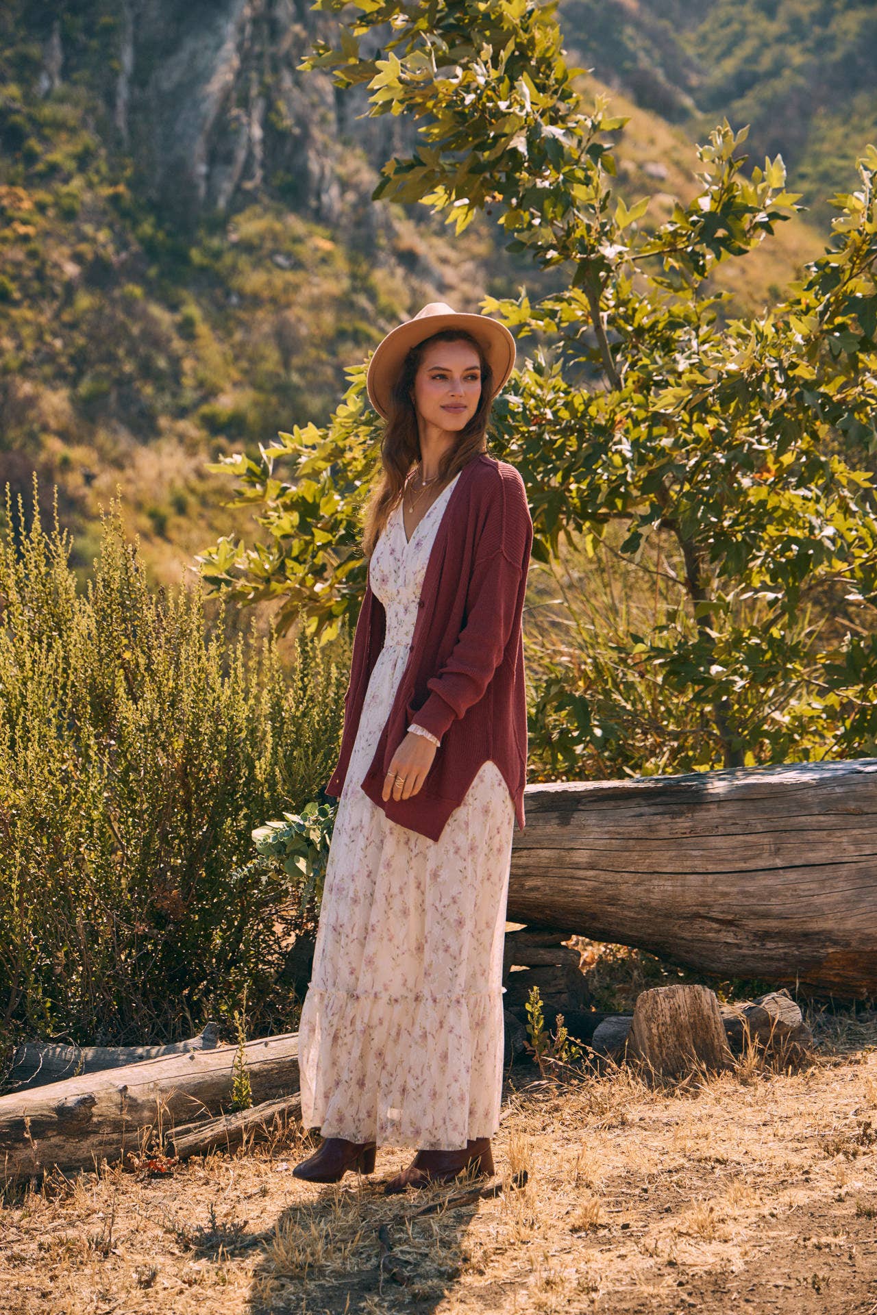 Front view — cream floral maxi dress with smocked waist and ruffle hem; mauve cardigan, tan hat, holding a greenery bouquet outdoors.