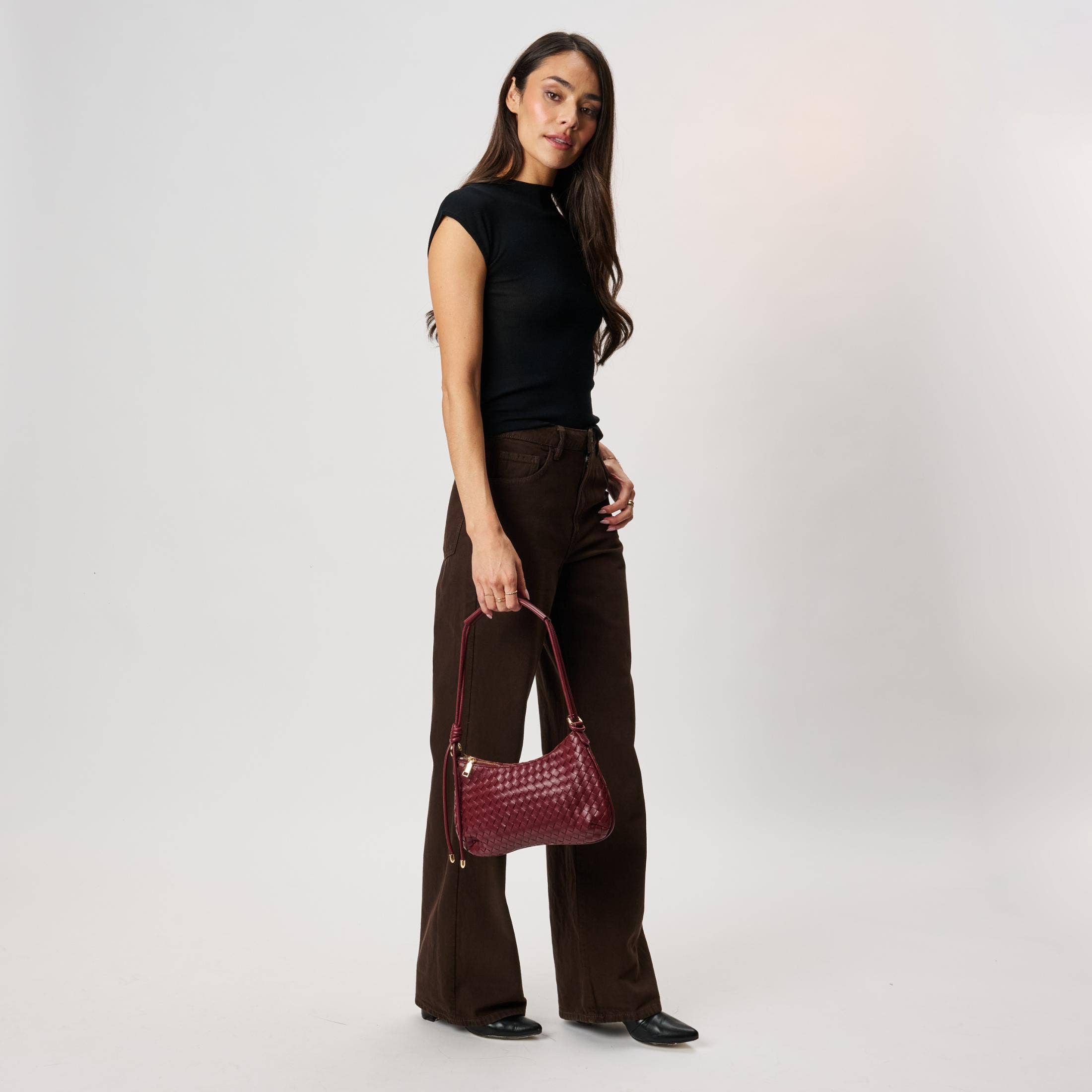Cindy Woven Crescent Shoulder Bag in Burgundy by Moda Luxe — model shoulder carry view showing woven vegan leather crescent bag with knotted strap, gold-tone hardware and zip closure, styled with black top and dark brown trousers