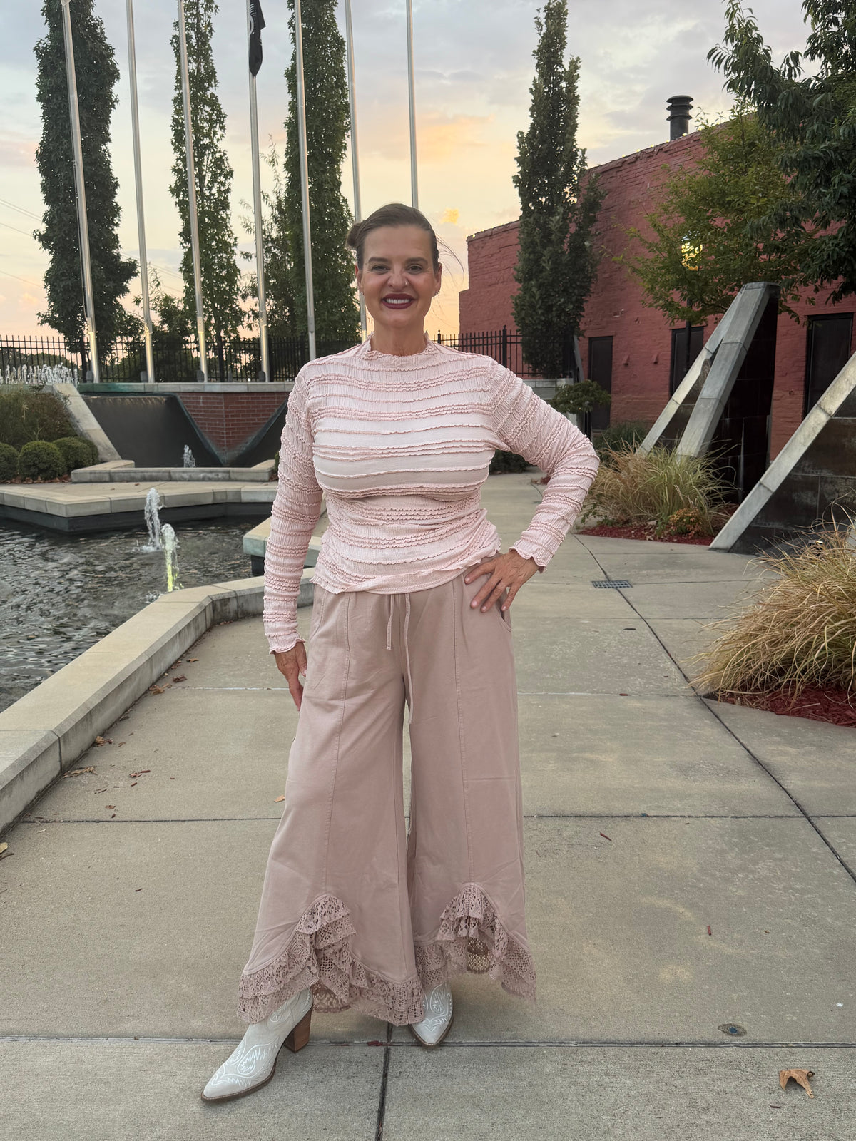 A woman stands outdoors on a paved walkway near a water feature and brick building, wearing a light pink ribbed long-sleeve top and matching wide-leg ruffled pants with white pointed boots.