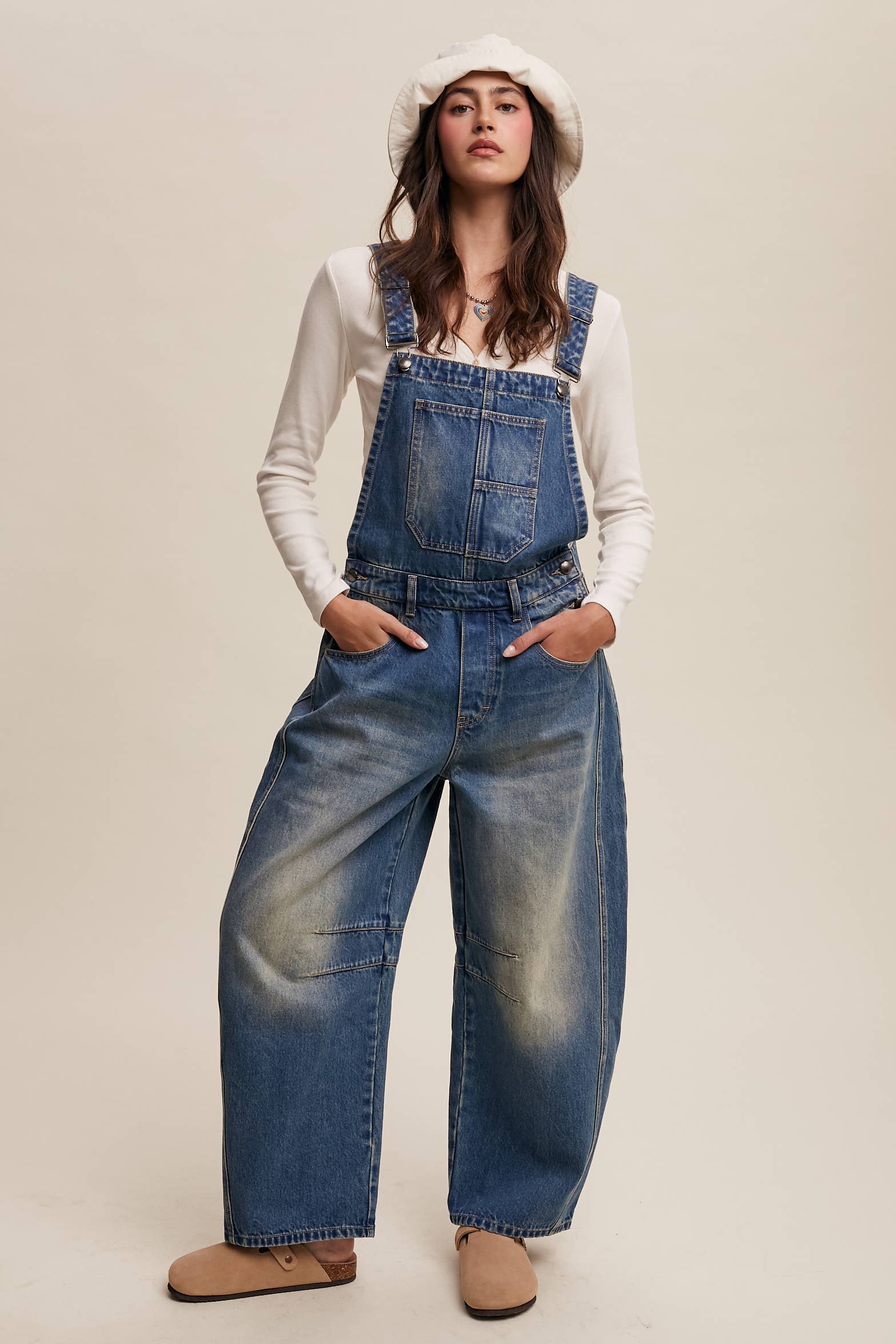 Vintage-inspired blue denim overalls with relaxed wide-leg fit, front chest pocket, and adjustable straps, styled with a white long-sleeve top and neutral clogs.