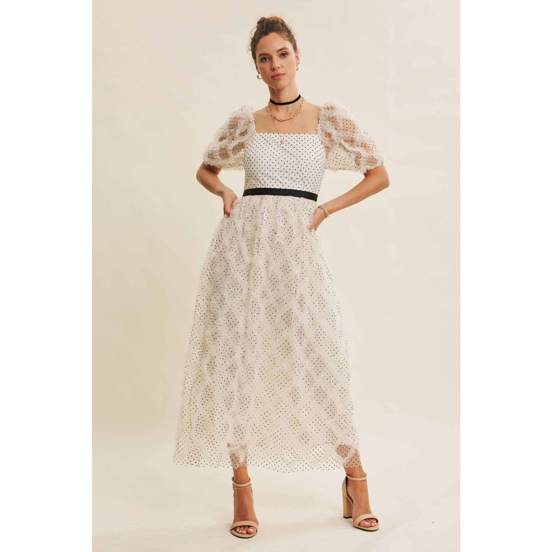 Front view — cream polka-dot tulle midi dress with puff sleeves and black belt; styled with black hat, ankle boots, pearl clutch; leaning by wooden doors.