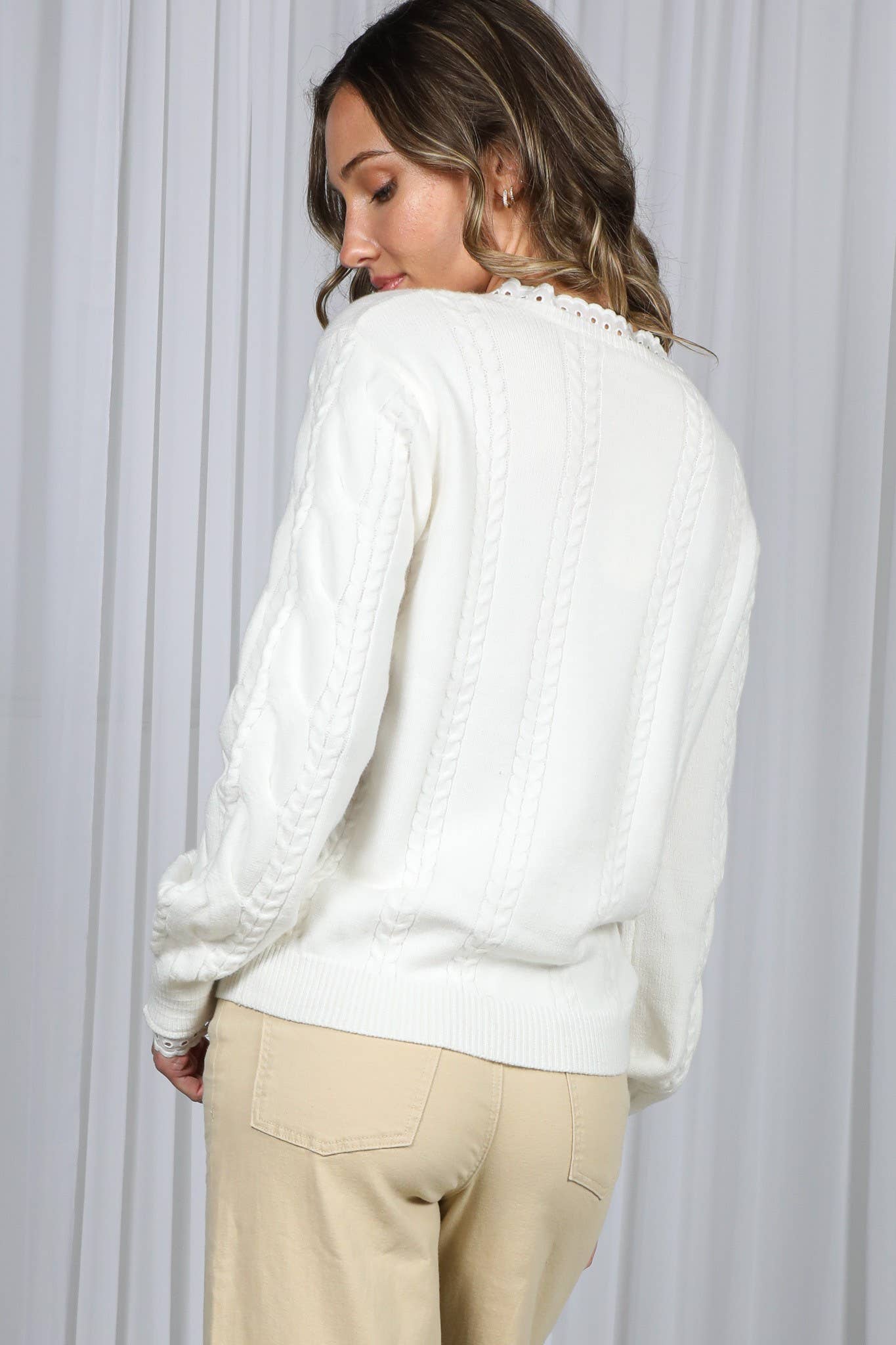 Back view Side view of Model wearing a off-white V-neck cable knit sweater with lace trim and ruffle detail, paired with a beige pants.