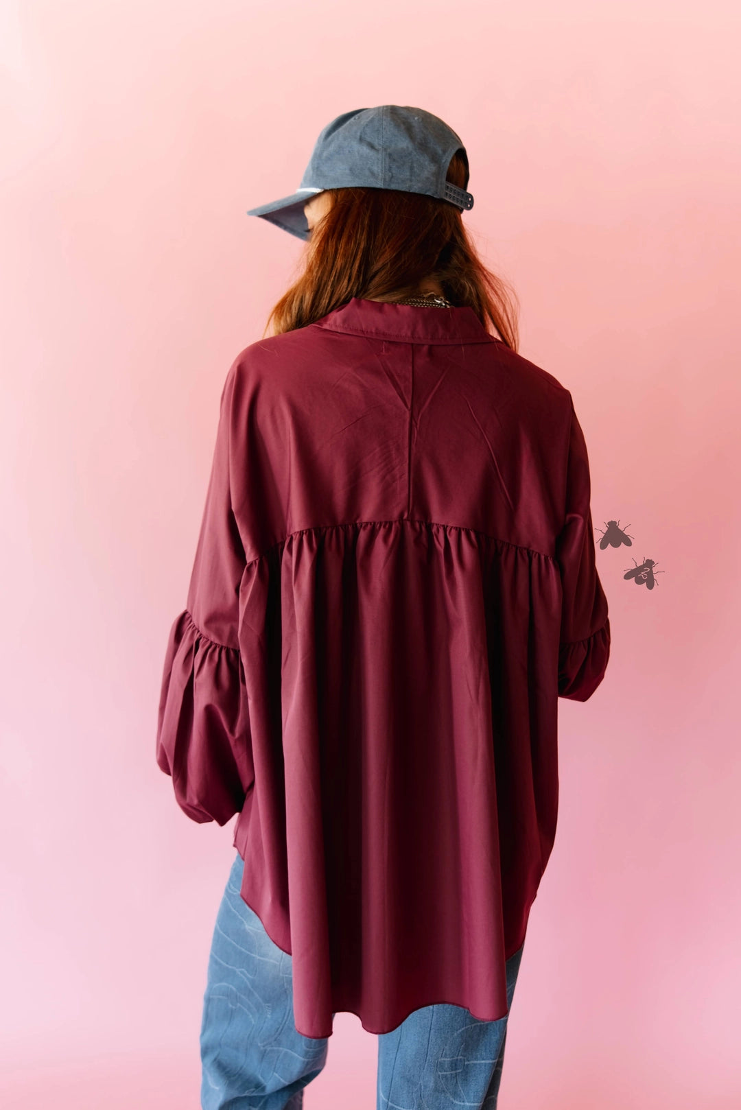 A person shown from the back wearing an oversized burgundy blouse with gathered sleeves, paired with blue jeans and a blue cap, set against a solid pink background.