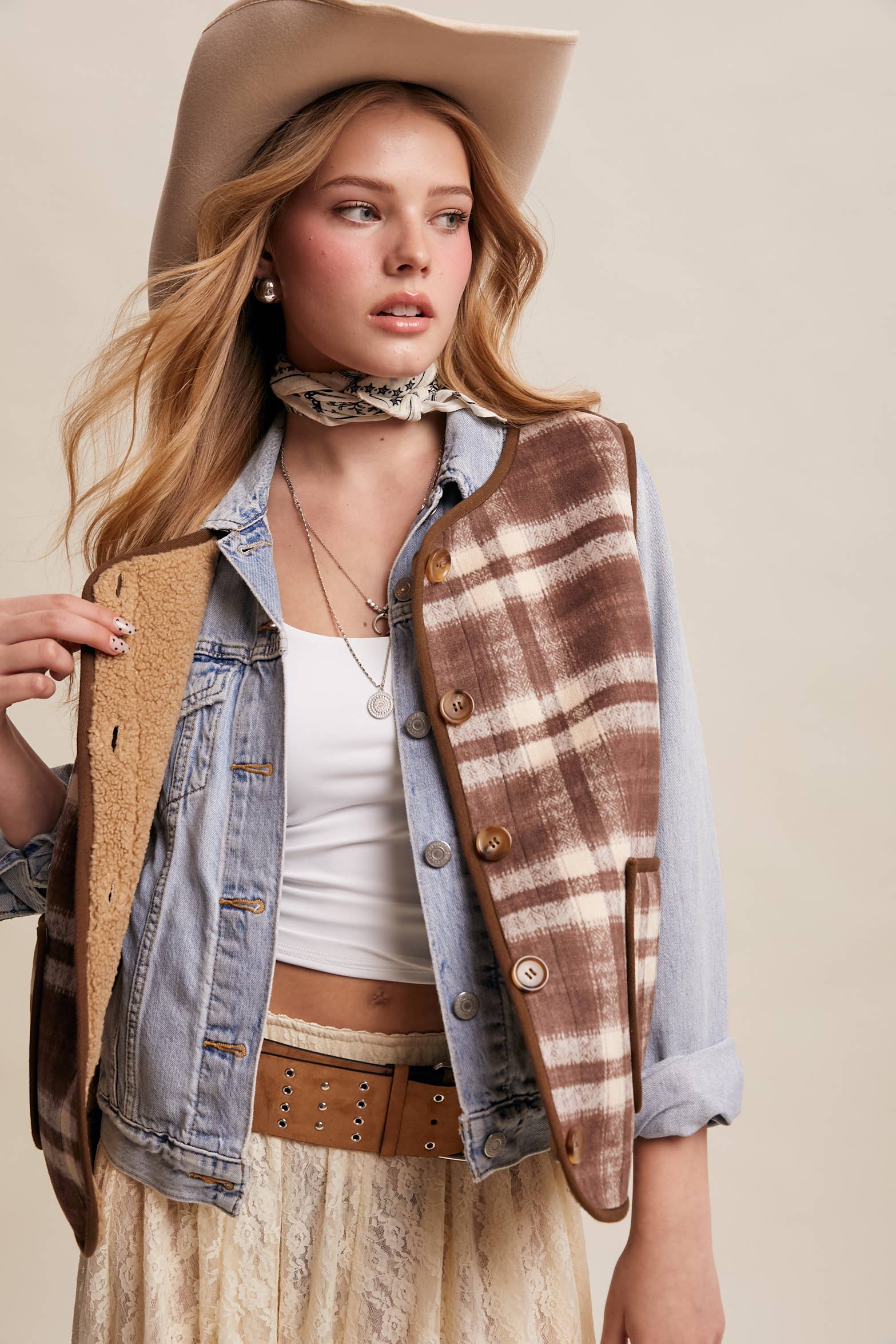 Woman wearing a plaid vest, denim jacket, and cowboy hat against a beige background