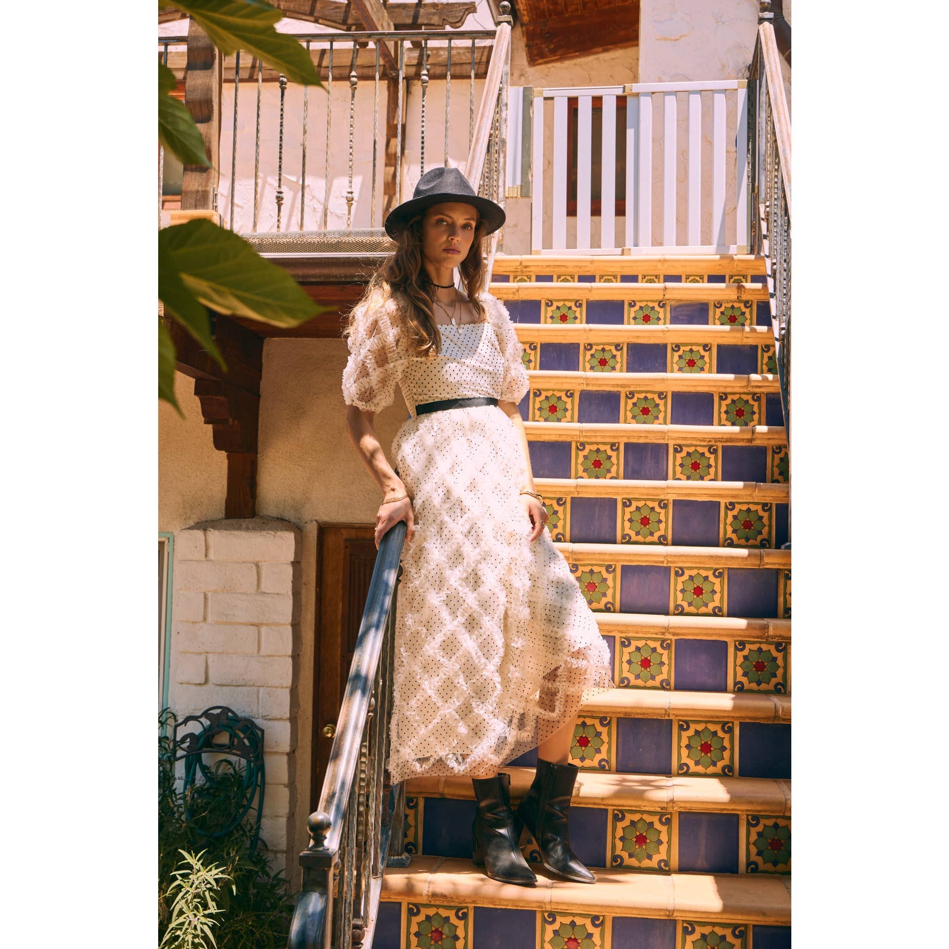 Front view — cream polka-dot tulle midi dress with puff sleeves and black belt; styled with black hat, ankle boots, pearl clutch; leaning by wooden doors.