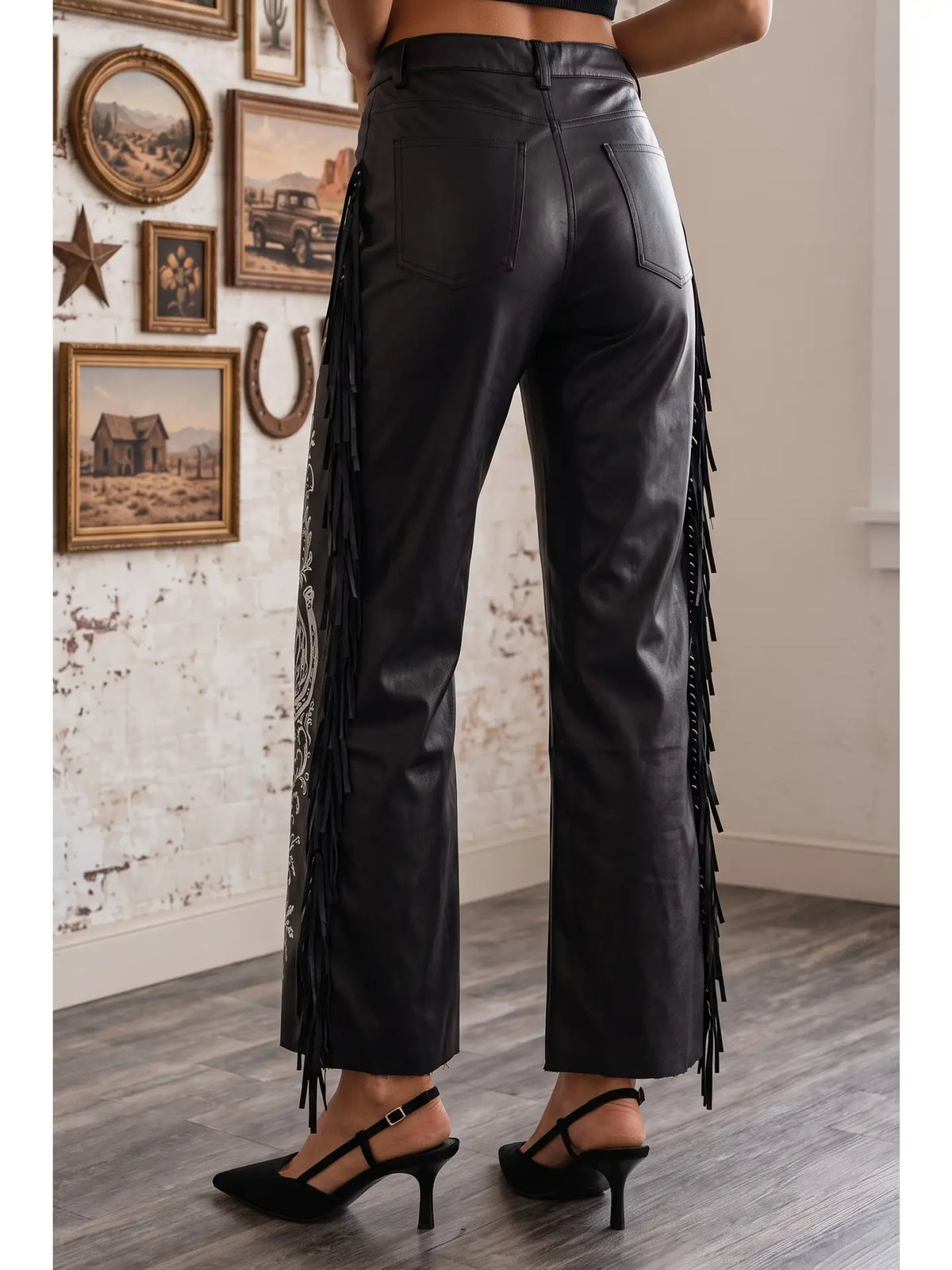 Black faux-leather cropped pants with long fringe detailing down the sides, shown on a model from the back in a western-themed room.