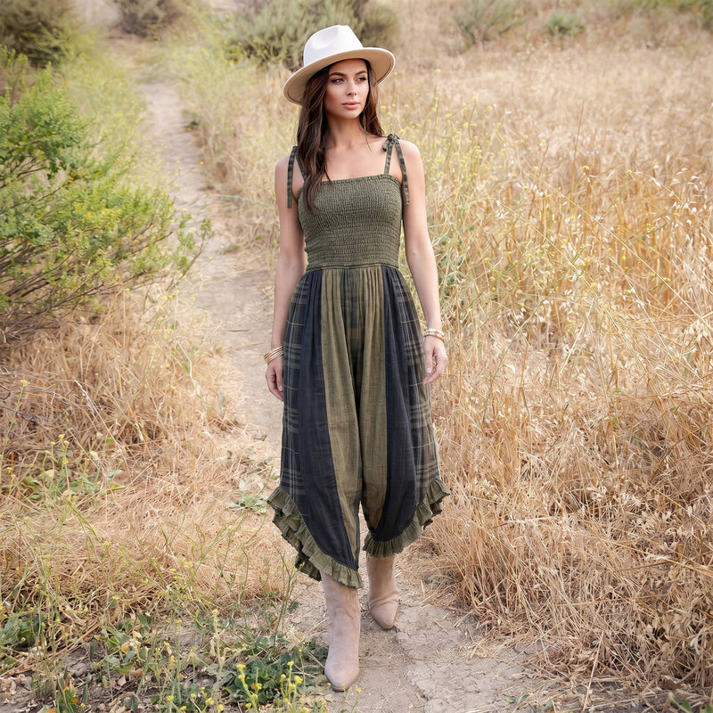 A woman stands on a dirt path in a dry grassy field wearing a sleeveless olive green and black plaid maxi dress with a textured bodice, a cream wide-brim hat, and beige ankle boots.