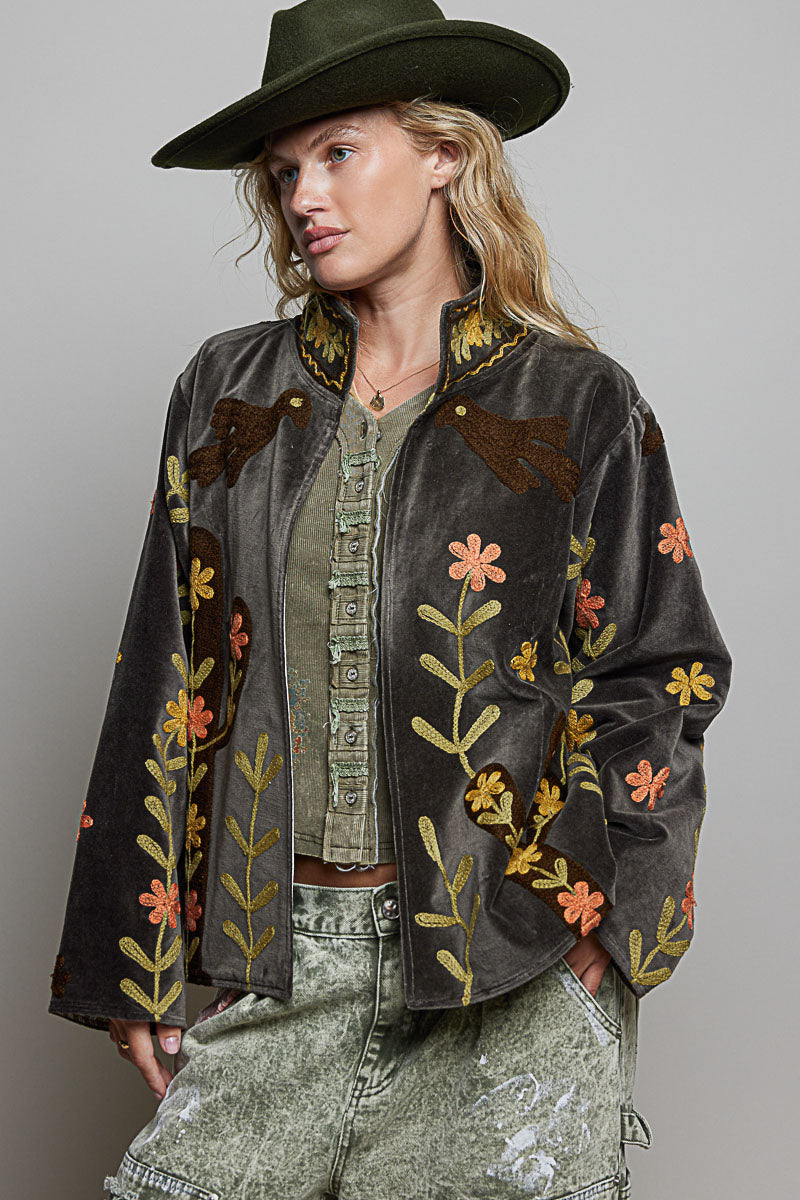 A woman wearing a dark brown velvet jacket with colorful floral embroidery, an olive green wide-brim hat, and distressed light green jeans, photographed against a neutral gray studio background.