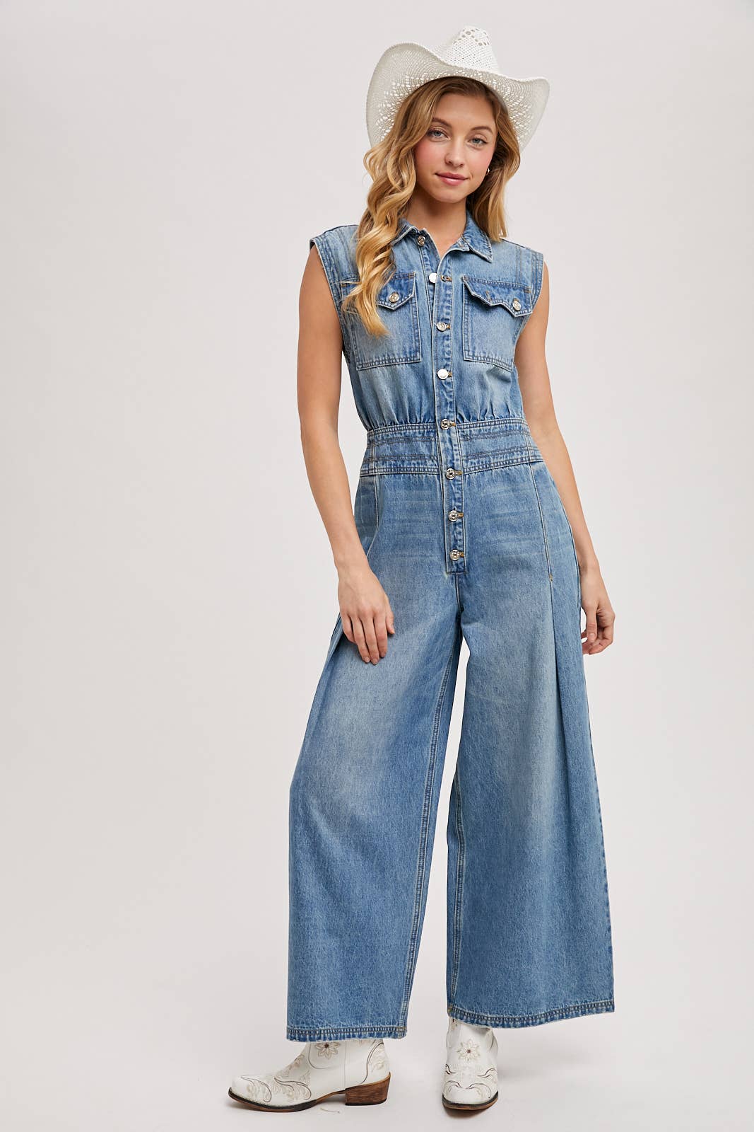 Sleeveless denim jumpsuit with collared button-front, chest flap pockets, and wide-leg palazzo silhouette, shown on model with slide sandals.