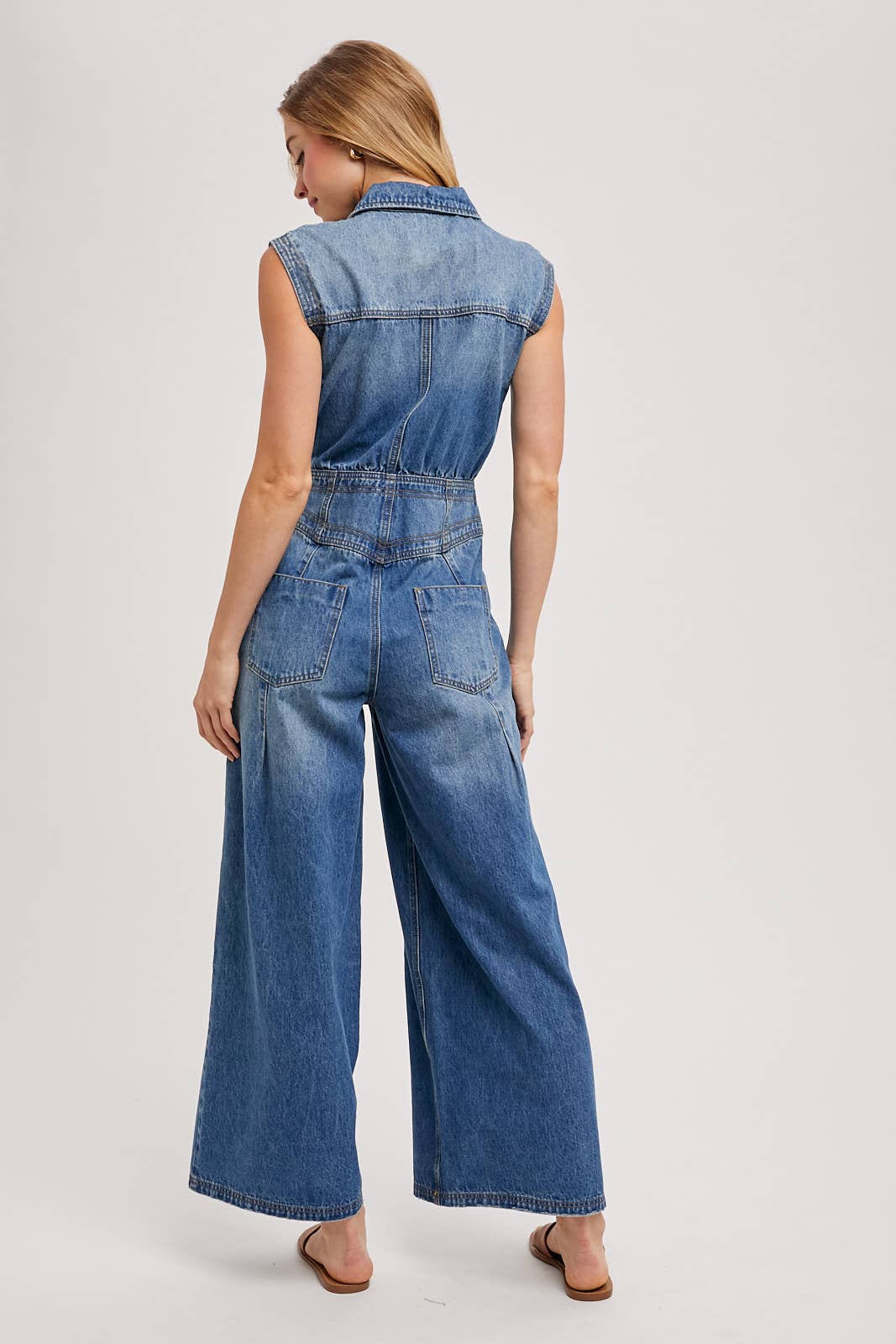 Back view of sleeveless denim jumpsuit with collared neckline, defined waist, back patch pockets, and wide-leg pants, worn with brown slide sandals.