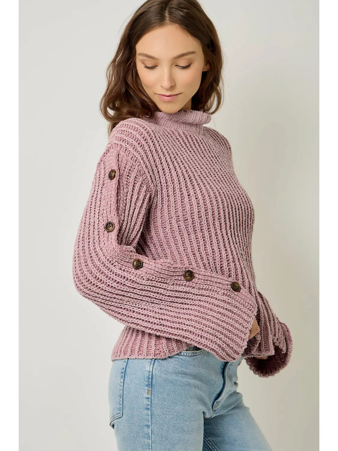 A woman wearing a chunky ribbed knit sweater in dusty mauve pink with dark brown buttons, paired with light blue jeans, against a plain light gray background.
