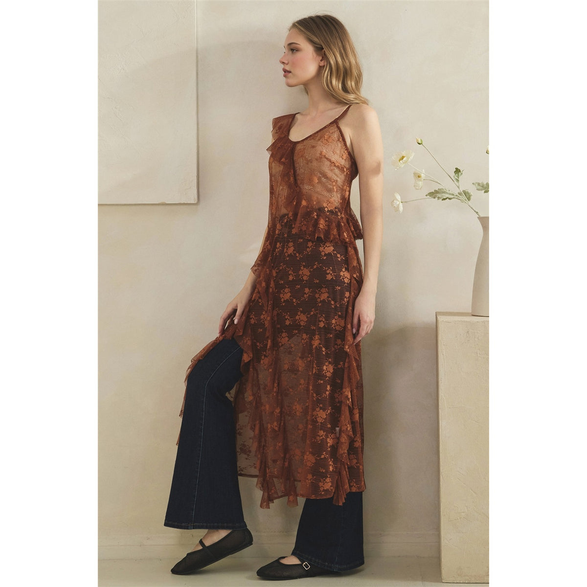 A woman models a copper sheer floral midi duster with spaghetti straps and a ruffled neckline, styled with dark wide-leg jeans and black flats in a minimalist beige interior setting.
