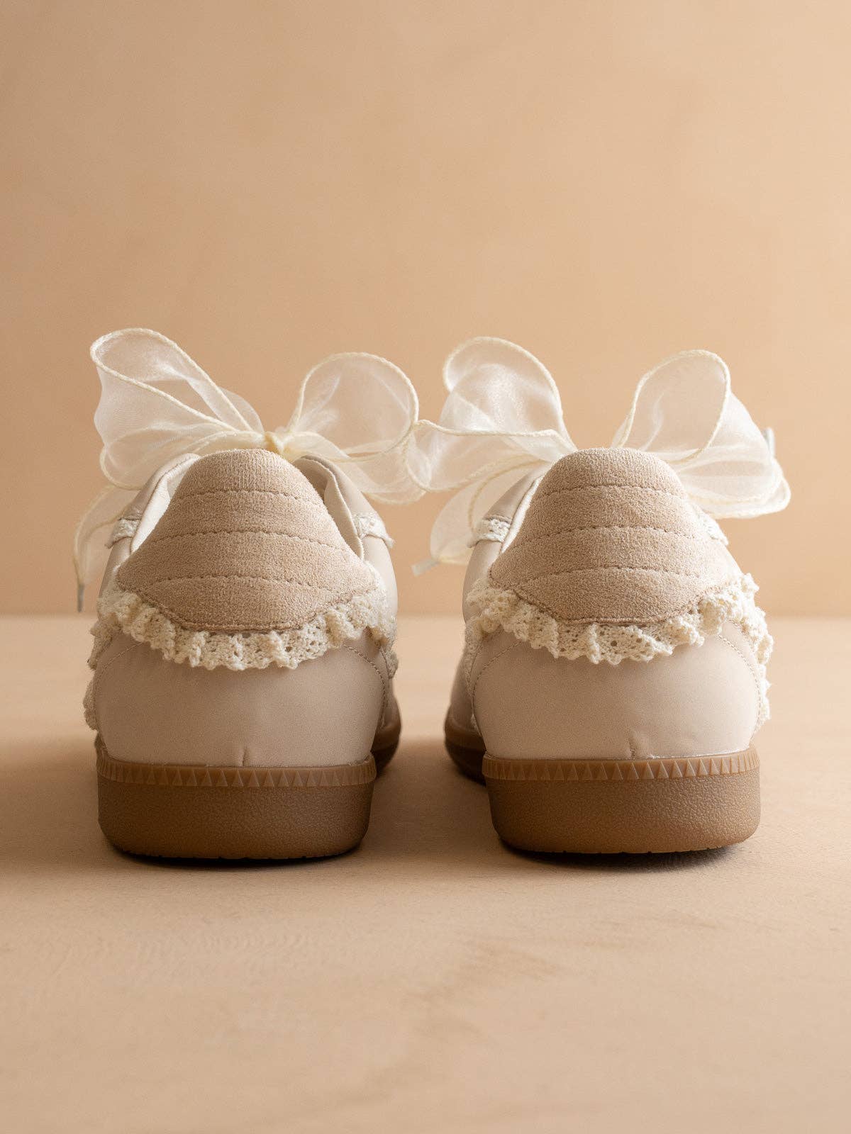 Back view of ivory lace-trim sneakers with oversized organza ribbon bows, quilted heel tabs, and gum soles on a neutral background.