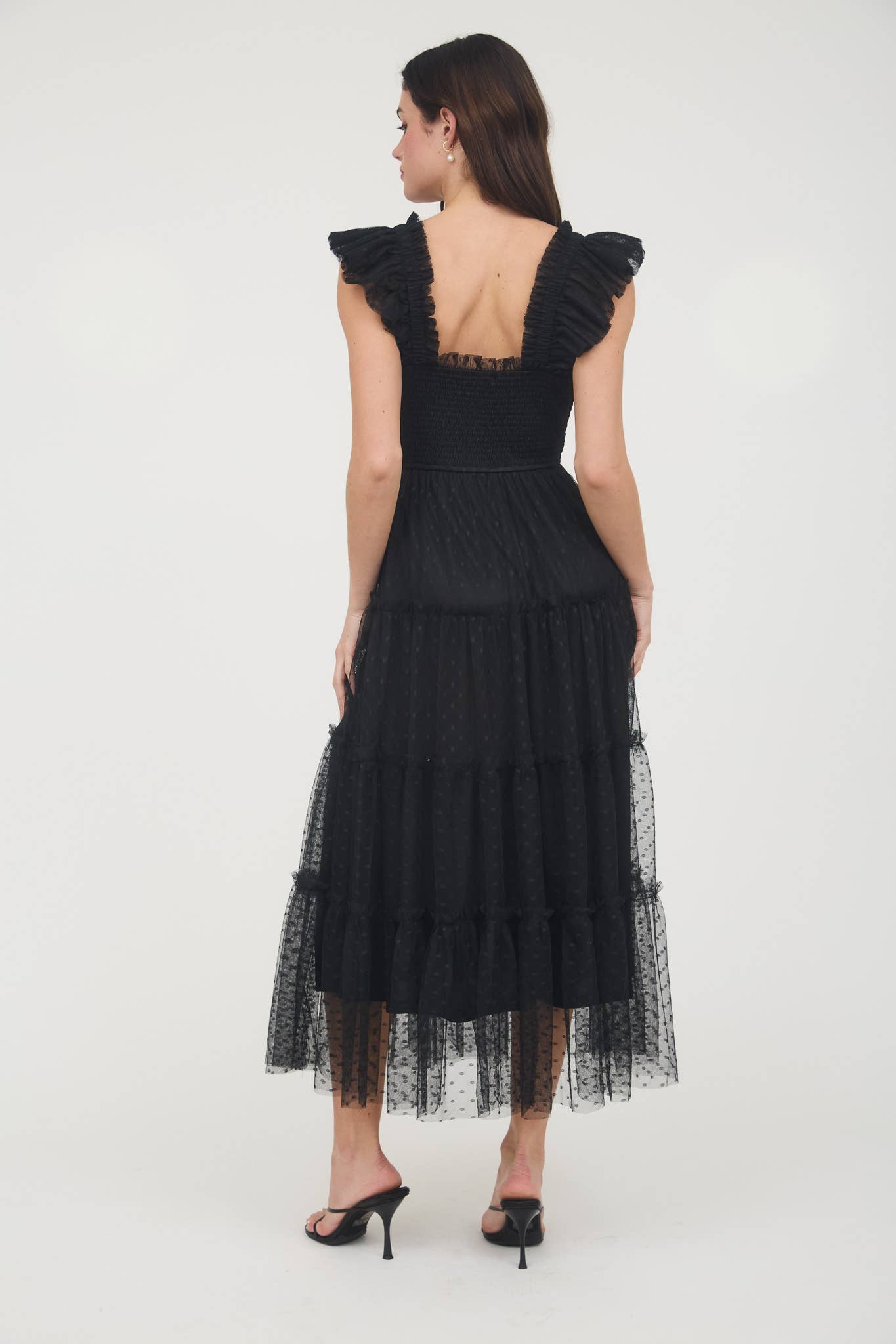 Back view of Model wearing black smocked midi dress with ruffle sleeves, tiered dotted mesh overlay, and flowy A-line silhouette.