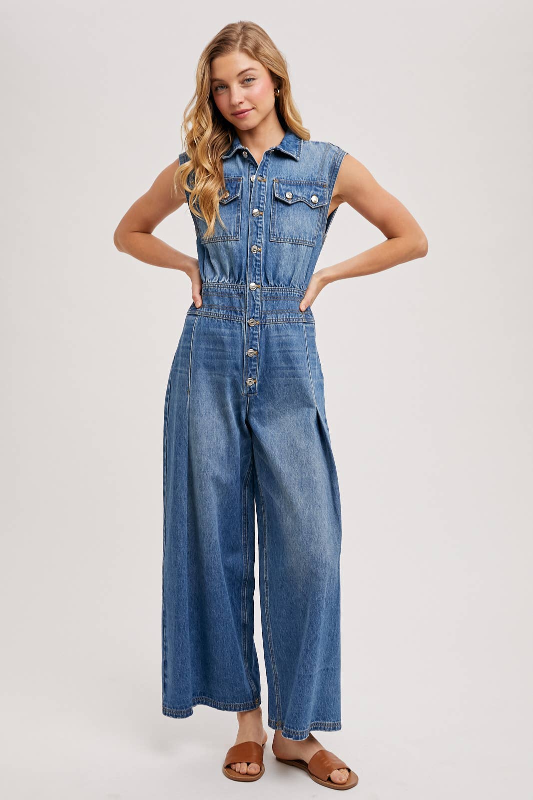 Sleeveless denim jumpsuit with collared button-front, chest flap pockets, and wide-leg palazzo silhouette, shown on model with slide sandals.