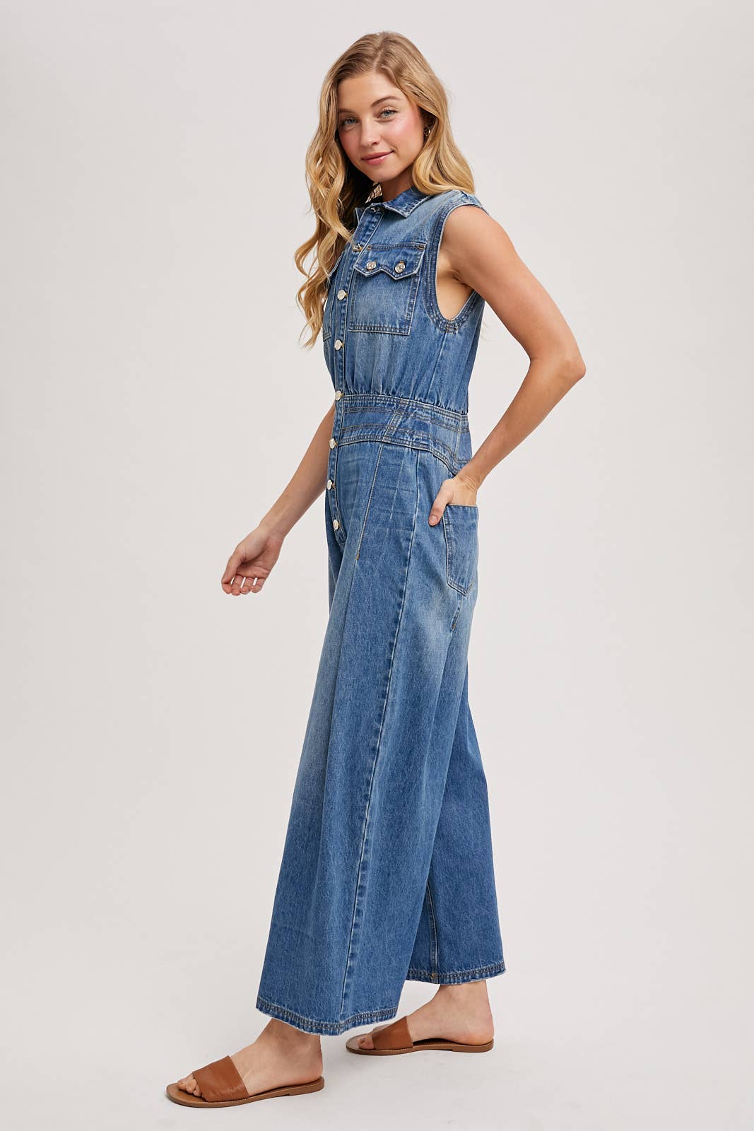 Side view of sleeveless denim jumpsuit with collared button-front and wide-leg legs, worn with brown slide sandals.