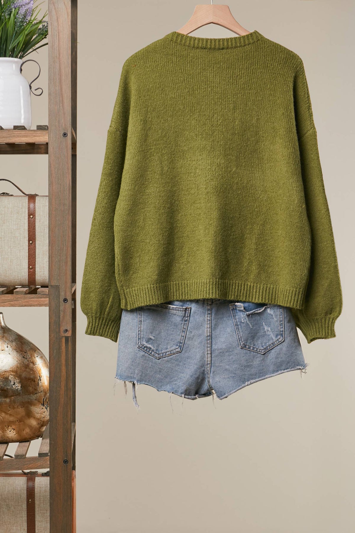 An olive green knit sweater hanging on a wooden hanger, styled with light-wash denim shorts against a beige wall, with a rustic wooden shelf visible on the left.