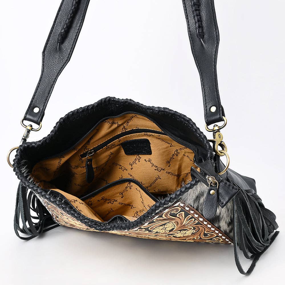A black leather shoulder bag with a western-style tooled leather panel, black and white cowhide, and black leather fringe tassels, shown open to display its tan patterned interior.
