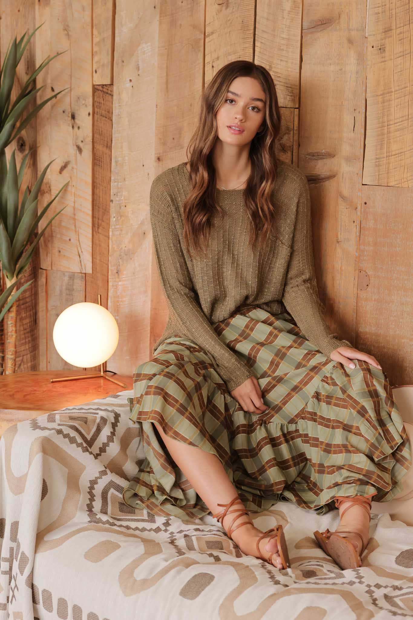 Model wearing olive distressed lightweight  distressed sweater with ribbed texture and front pocket, paired with a plaid ruffle skirt in rustic tones