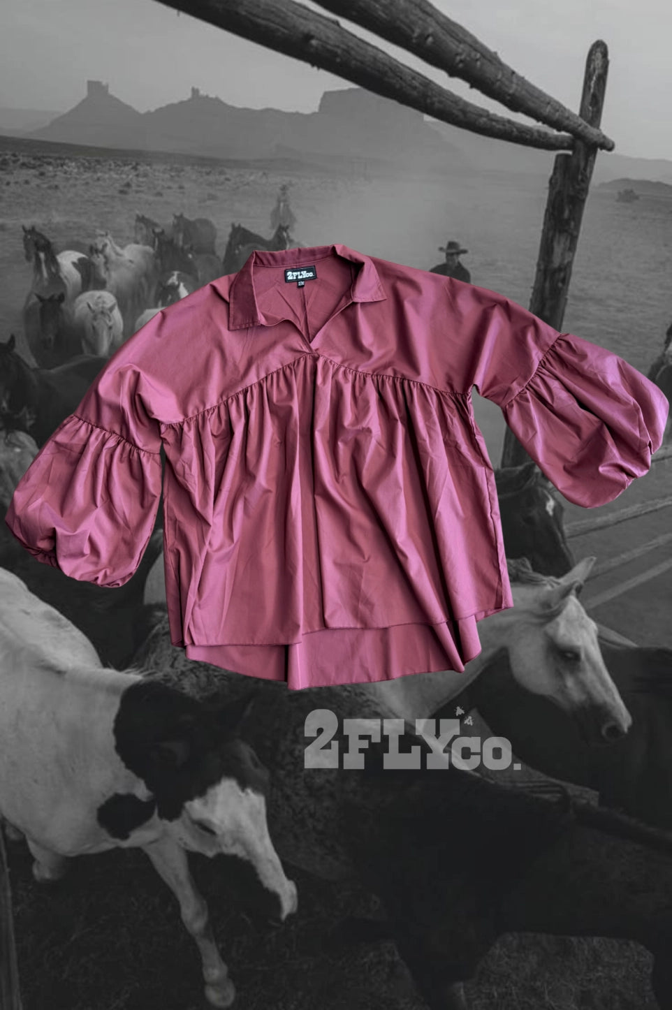 A dusty rose pink long-sleeve blouse with a ruffled yoke and gathered sleeves, shown against a black and white western ranch background with cows and a wooden fence. The brand name '2FLYCo.
