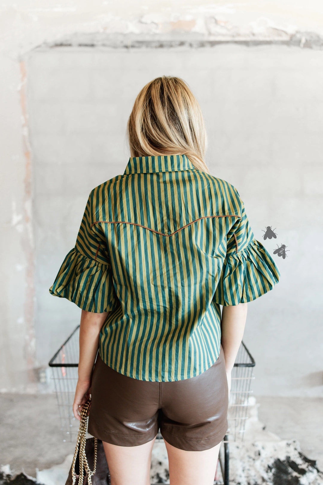A woman viewed from behind wearing a green and yellow vertically striped western-style blouse with ruffled sleeves and brown shorts, standing in front of a textured concrete wall.