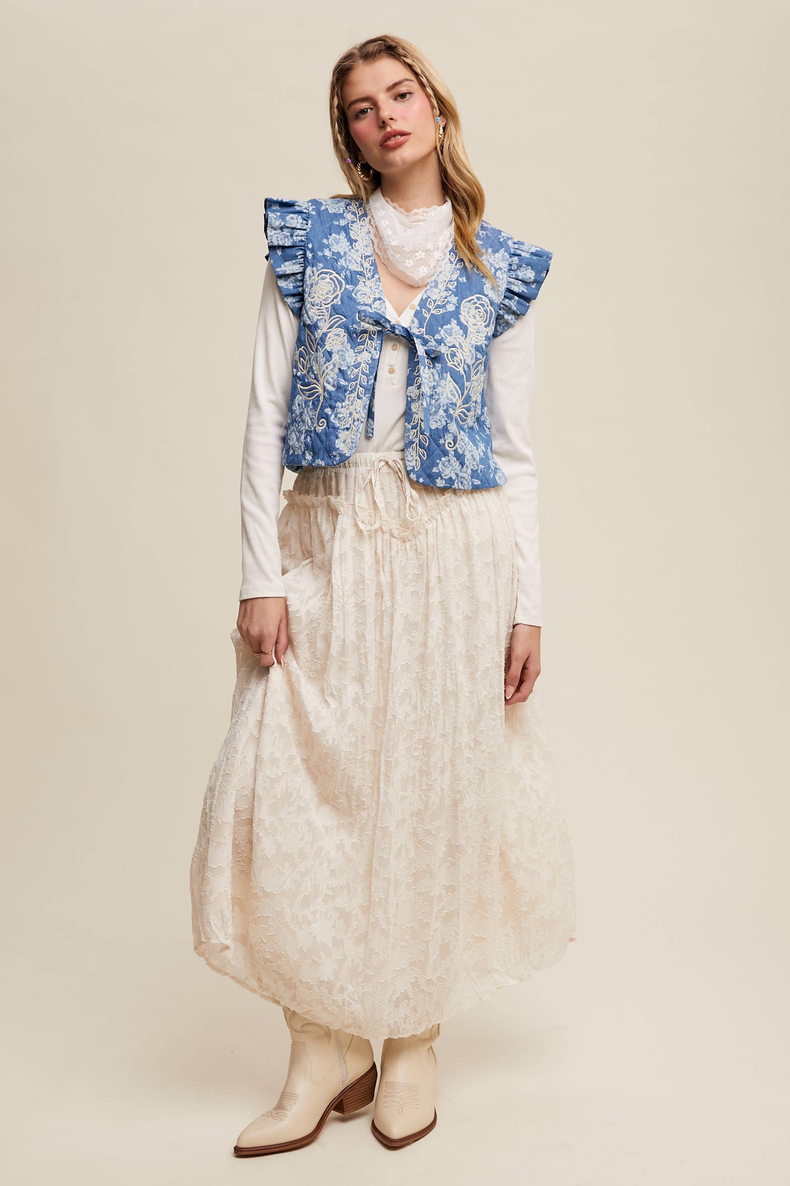 Ivory floral lace maxi skirt with drawstring waist and bubble hem, styled with a white long-sleeve top, blue ruffled embroidered vest, and cream cowboy boots.