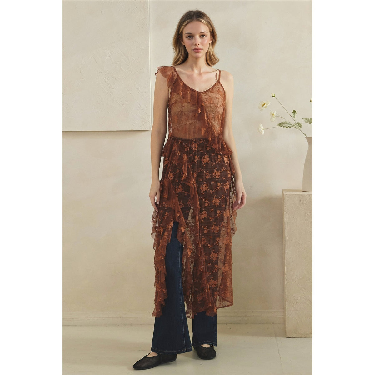 A woman models a copper sheer sleeveless tunic with a floral pattern and layered ruffle hem, styled over dark blue jeans and black flats in a minimalist beige interior setting.