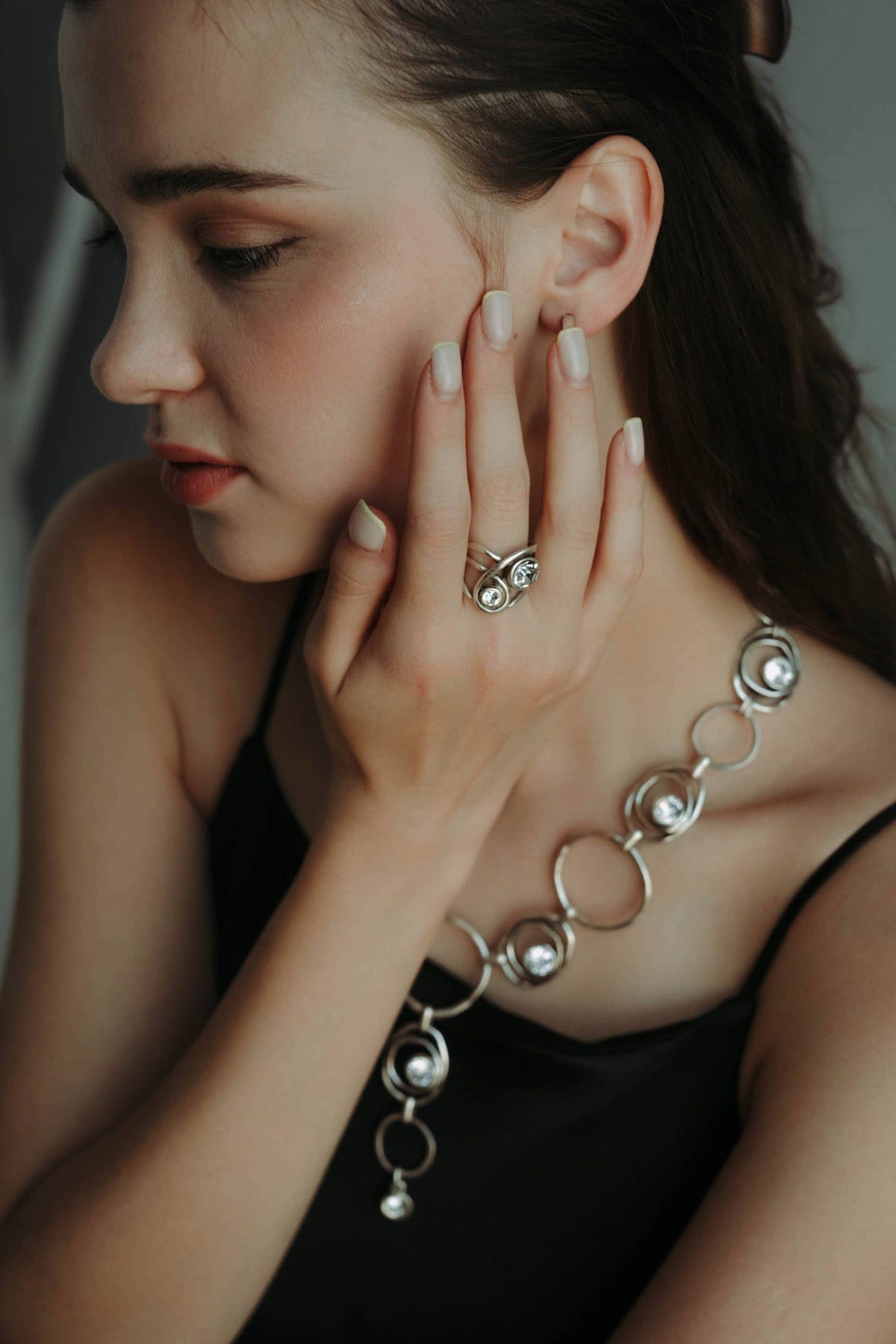 A close-up lifestyle shot of a woman wearing a silver-toned geometric ring on her finger and a matching silver chain necklace with circular links and pearl-like beads, paired with a black top.
