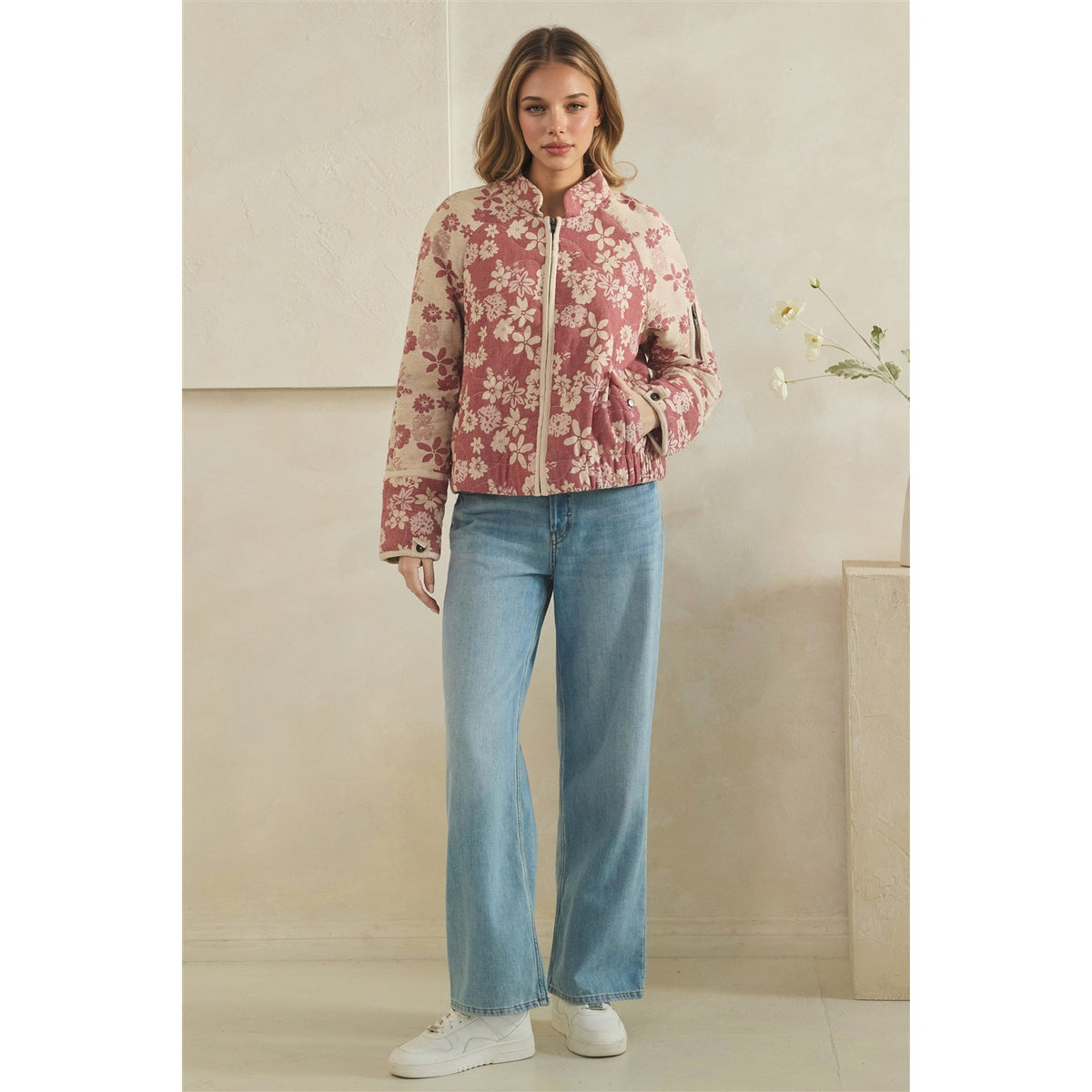 A woman stands indoors wearing a cropped floral jacket in dusty pink and cream, light blue jeans, and white sneakers. The background is a warm beige wall with a small decorative plant and pedestal.