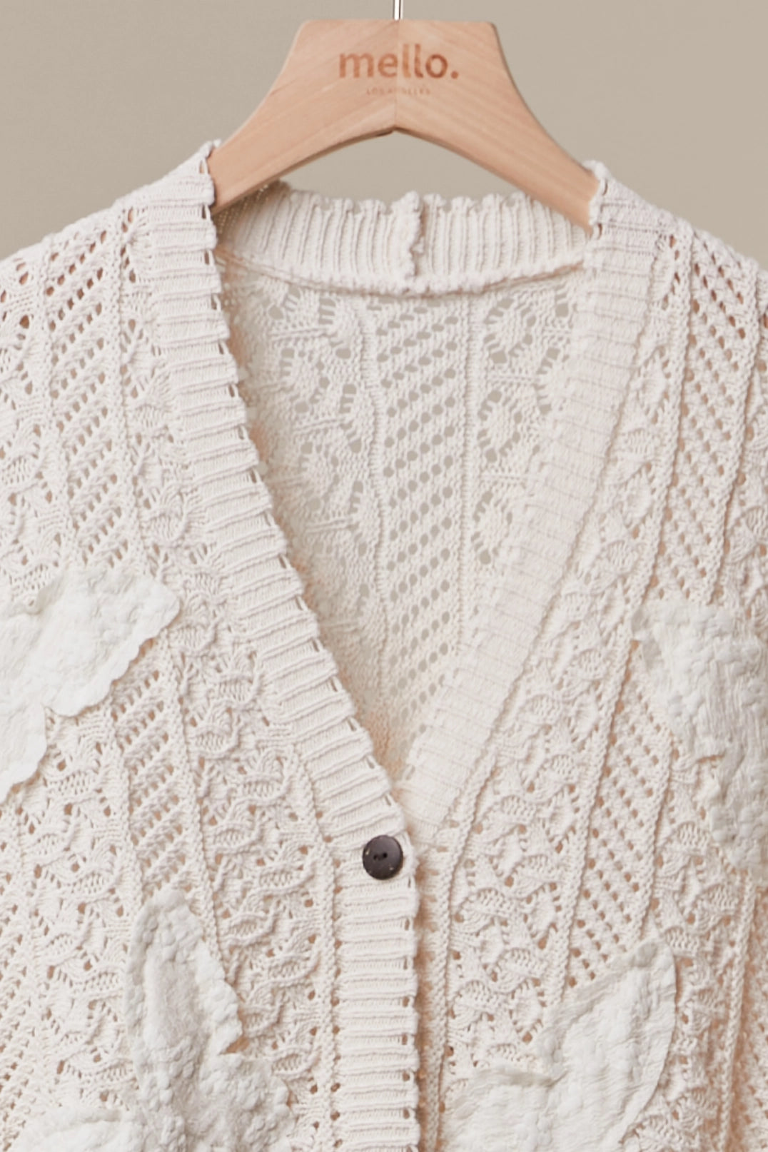 An off-white textured knit cardigan sweater with lace and floral patterns, hanging on a wooden hanger branded 'mello.' against a neutral beige background.
