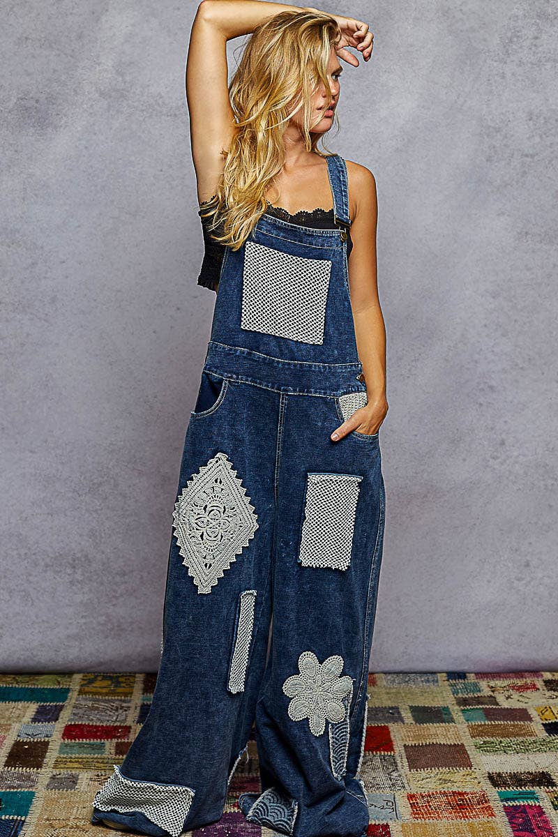 Boho denim patchwork overalls featuring wide legs, crochet lace appliqué patches, and vintage-inspired distressed detailing. Styled with a black crop top and sneakers for a relaxed, eclectic look.