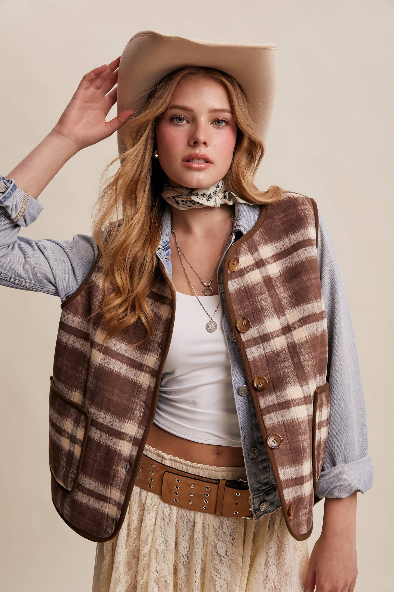 Woman wearing a plaid vest, cowboy hat, and lace skirt against a beige background