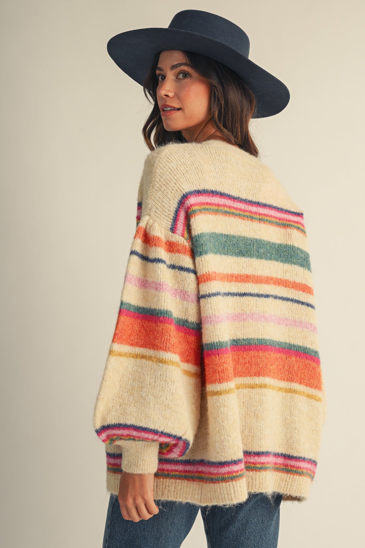 A woman wearing a cream-colored knit poncho sweater with colorful horizontal stripes in pink, orange, green, and blue, paired with a dark navy wide-brim hat and blue jeans, photographed against a solid light beige background.
