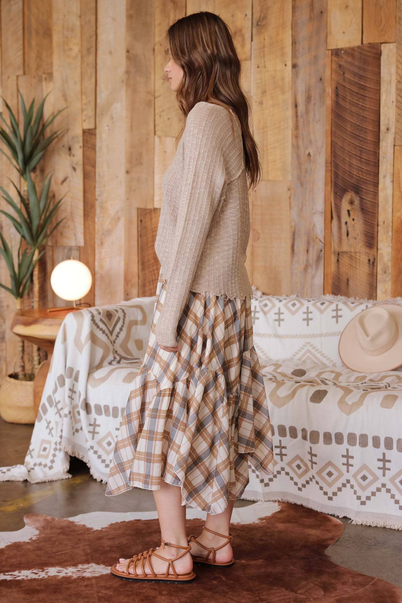 Side view of model wearing taupe distressed lightweight sweater with ribbed knit texture, raw edge hem, and relaxed fit paired with tan plaid ruffle skirt