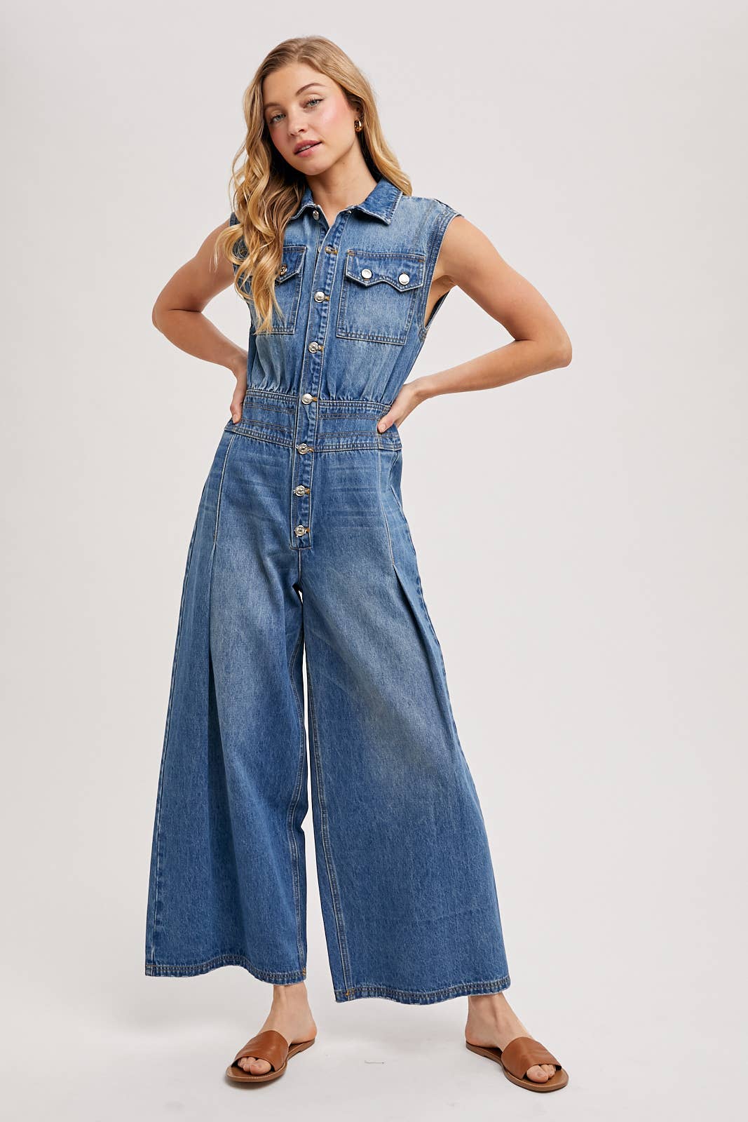 Sleeveless denim jumpsuit with collared button-front, chest flap pockets, and wide-leg palazzo silhouette, shown on model with slide sandals.