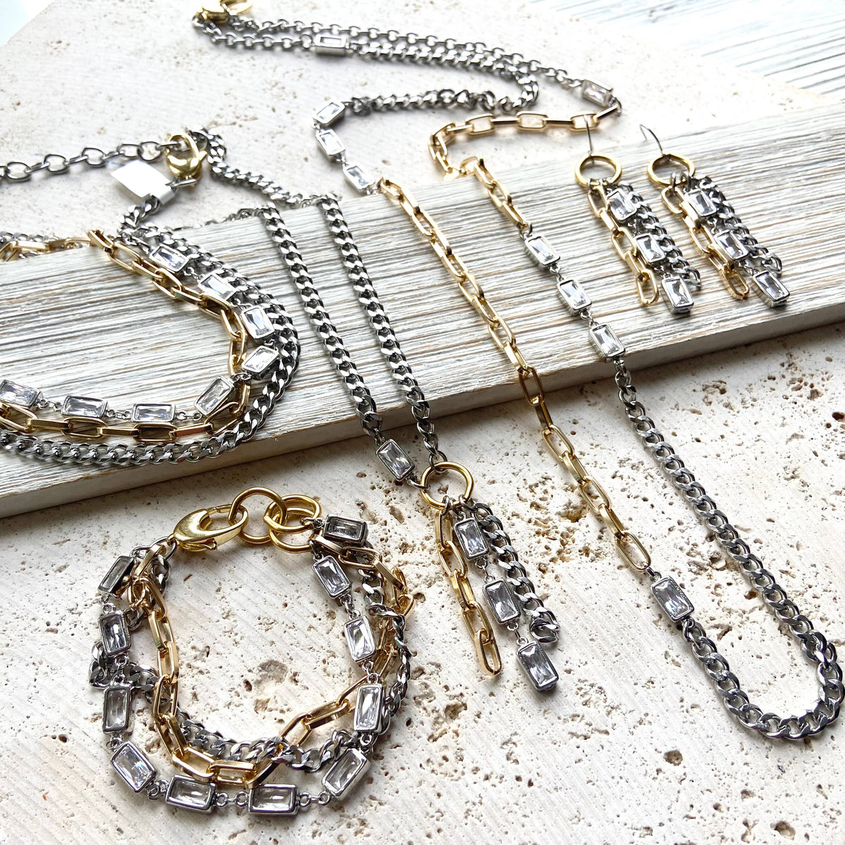 A styled flat-lay of a two-tone silver and gold jewelry set featuring a necklace, bracelet, and earrings, each adorned with clear baguette crystals, arranged on a distressed white wooden board over a textured beige surface.
