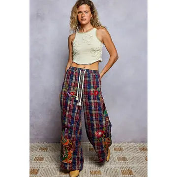 A woman modeling a pair of wide-leg plaid pants with colorful floral embroidery and a white drawstring, paired with a cream crop top, standing against a textured gray wall.