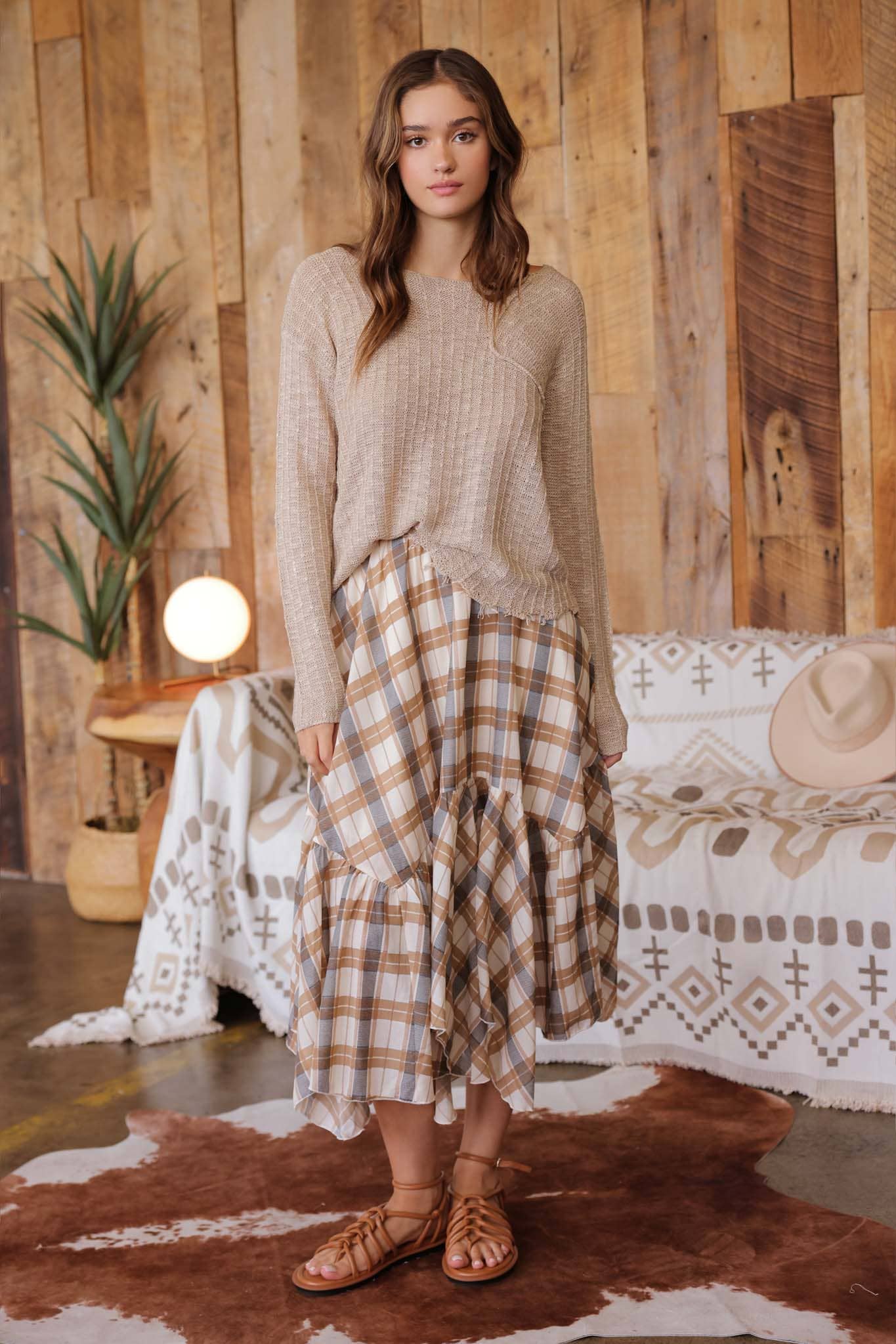 Woman wearing a beige sweater and plaid skirt standing in a rustic room with wooden walls and a patterned rug.